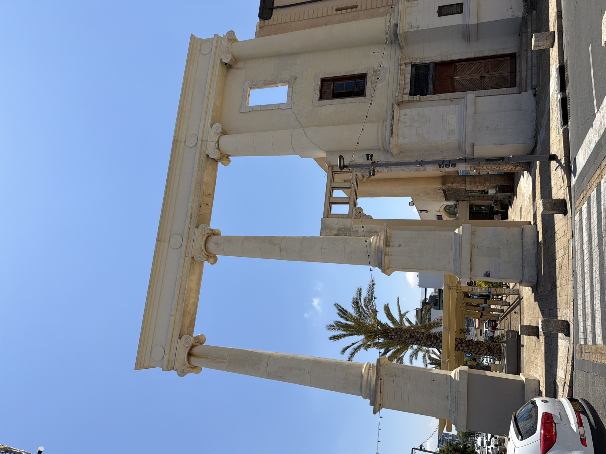 The image shows a low-angle, rotated view of the historical Jaffa Clock Tower in Tel Aviv, Israel, against a clear blue sky. The tower consists of three prominent columns supporting a structure with architectural details, including decorative elements and small windows. A palm tree is visible between the columns, adding a natural element to the urban setting. The street scene in the foreground includes part of a crosswalk, a parked white car, and glimpses of urban infrastructure, suggesting a busy city environment. The photograph captures the tower from an unusual angle, emphasizing its vertical structure and architectural design.