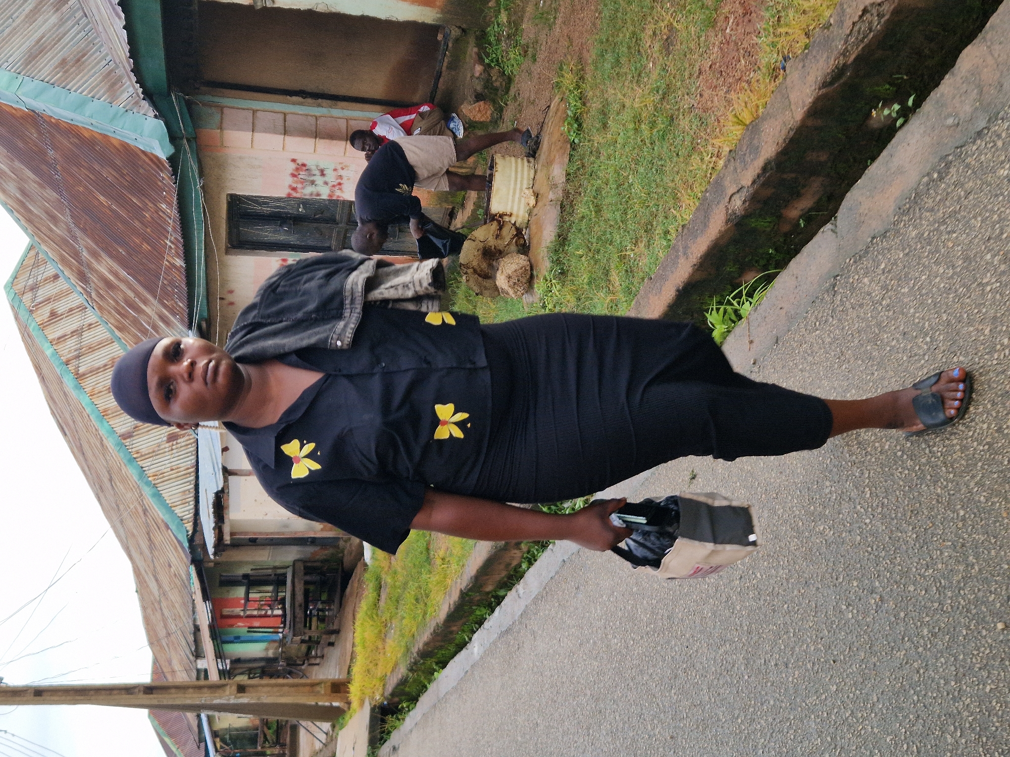A woman walks on a paved road in Kabong, Nigeria. She is wearing a black headscarf, a black short-sleeved shirt with yellow bow accents, and a long black skirt. She carries a beige and grey bag in her left hand. A dark jacket is draped over her shoulders. To the right of the road, a man in a black shirt and light brown shorts is bending over a metal drum, holding a black bag. Another person, wearing a red and white top, is seated behind him. The background features a row of buildings with corrugated metal roofs and colorful facades, with overgrown grass and a drainage ditch along the roadside.