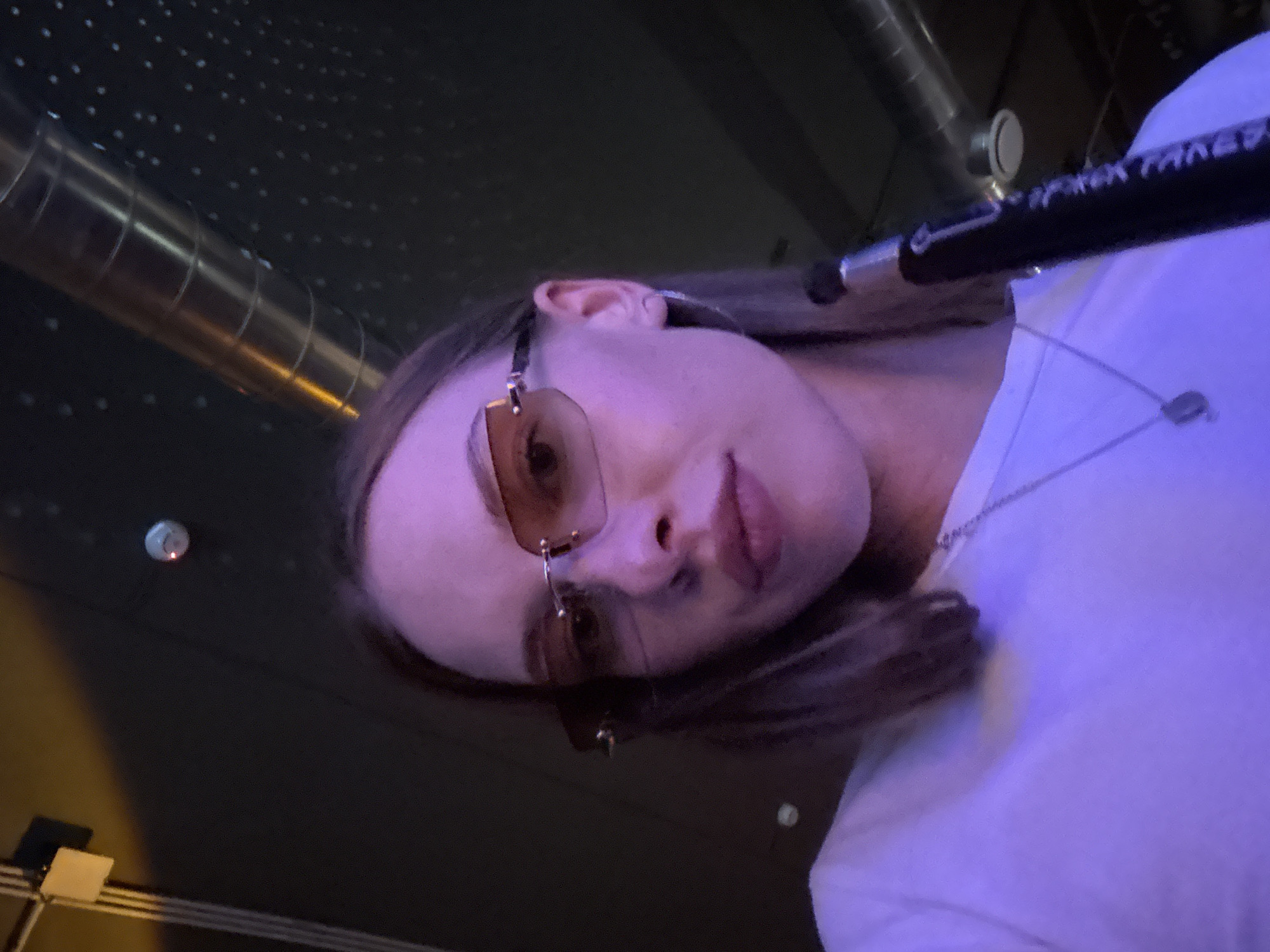 A young woman with shoulder-length brown hair and wearing glasses and a white t-shirt is captured in a selfie-style shot. She is wearing a silver necklace with a square pendant and a hoop earring in her left ear. A portion of a black object, possibly a hookah hose, is visible in the lower right of the frame. The environment appears to be dimly lit, with purple and yellow lighting casting a glow on the scene. The background shows industrial-style elements, including metallic pipes and what appears to be a ceiling. The scene is indoors, and there is no indication of the specific location within the city of Perm, Russia.