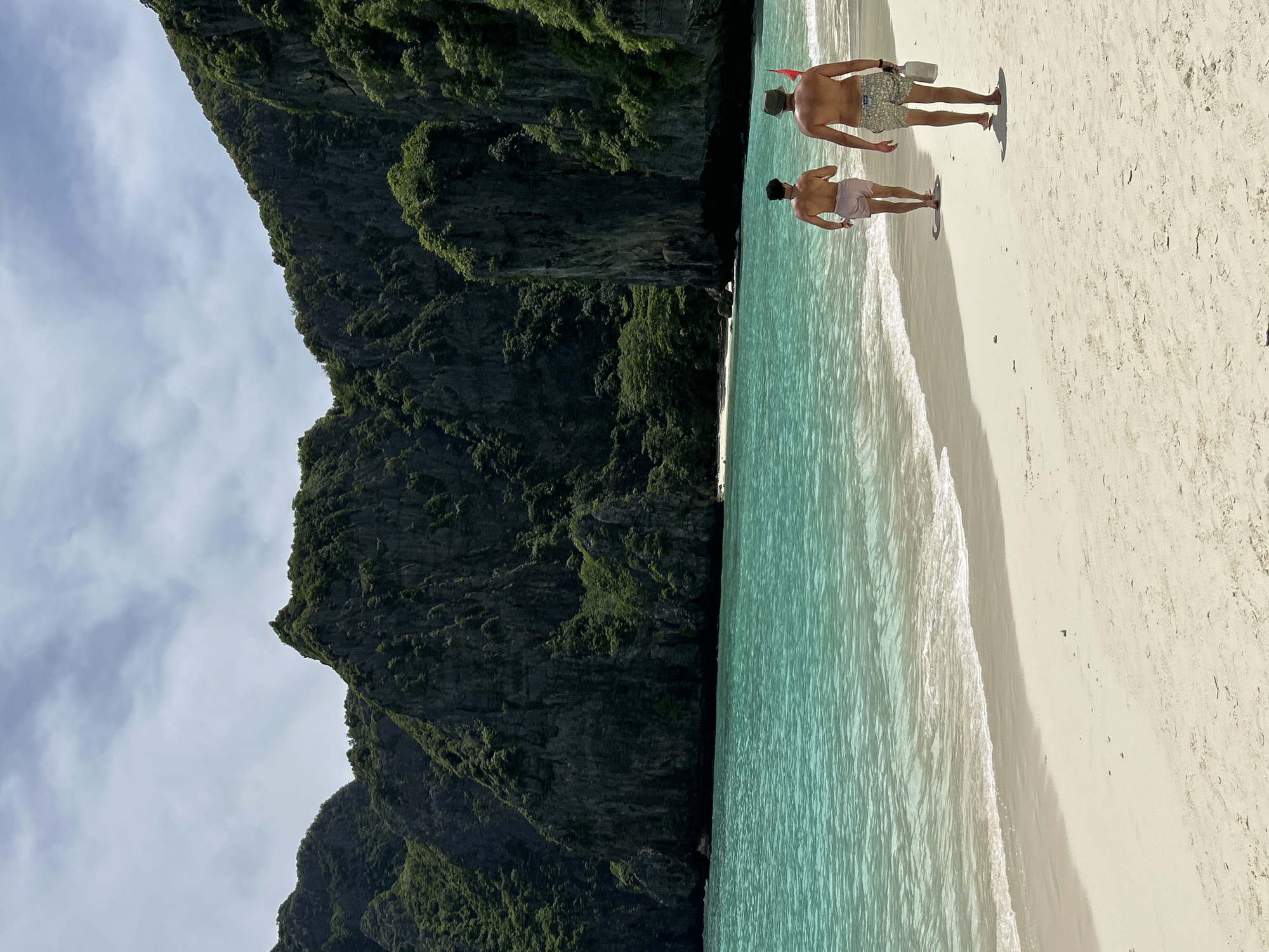 In Ao Nang, Thailand, two shirtless men in swim trunks walk along a sandy beach. The man on the left wears light pink shorts, while the man on the right has on patterned shorts and a hat. The man on the right carries a white bottle. The turquoise ocean laps at the shore, and behind it, dramatic, lush green cliffs rise under a partly cloudy sky.