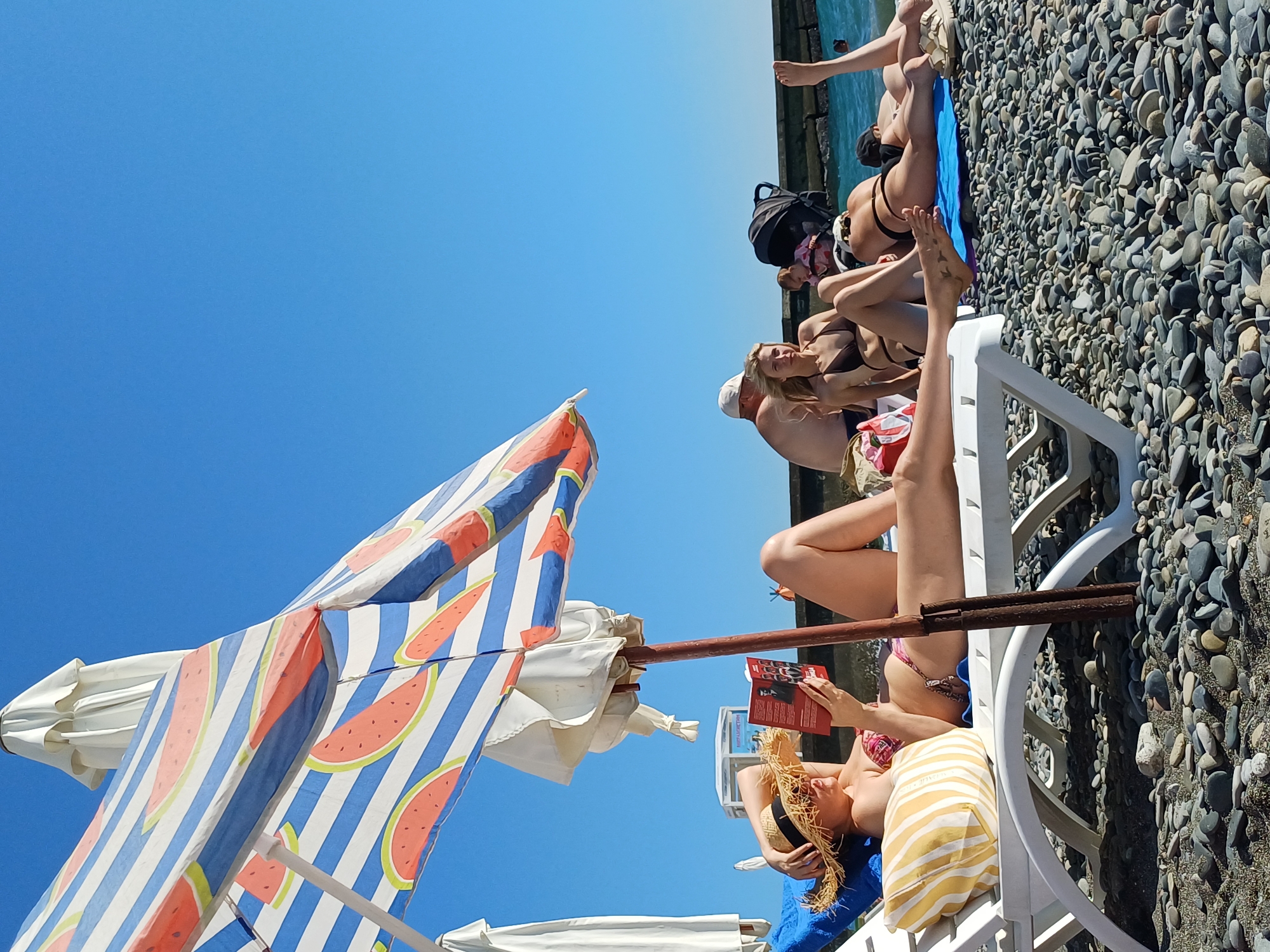 In a sun-drenched scene on a pebble beach in Sochi, Russia, several people are enjoying a day by the sea. The sky is a clear, bright blue, indicating fair weather. The focus of the image is a group of individuals lounging on beach chairs. One woman, wearing a straw hat and a bikini, is engrossed in a red book. Another person is partially visible, also in swimwear, with their legs extended. Behind them, a beach umbrella with a watermelon pattern provides shade, its vibrant stripes a contrast to the natural tones of the beach. Further back, a black backpack rests on the ground near the water's edge, suggesting a day of relaxation and leisure. The sea is calm, with a few people visible in the water in the distance. The overall mood is one of carefree enjoyment and summer vacation. The prominent objects are beach chairs, an umbrella, a backpack, and a book. The setting is distinctly a beach, characterized by the pebbles and the proximity to the water. The time of day appears to be midday, given the bright sunlight and clear skies.