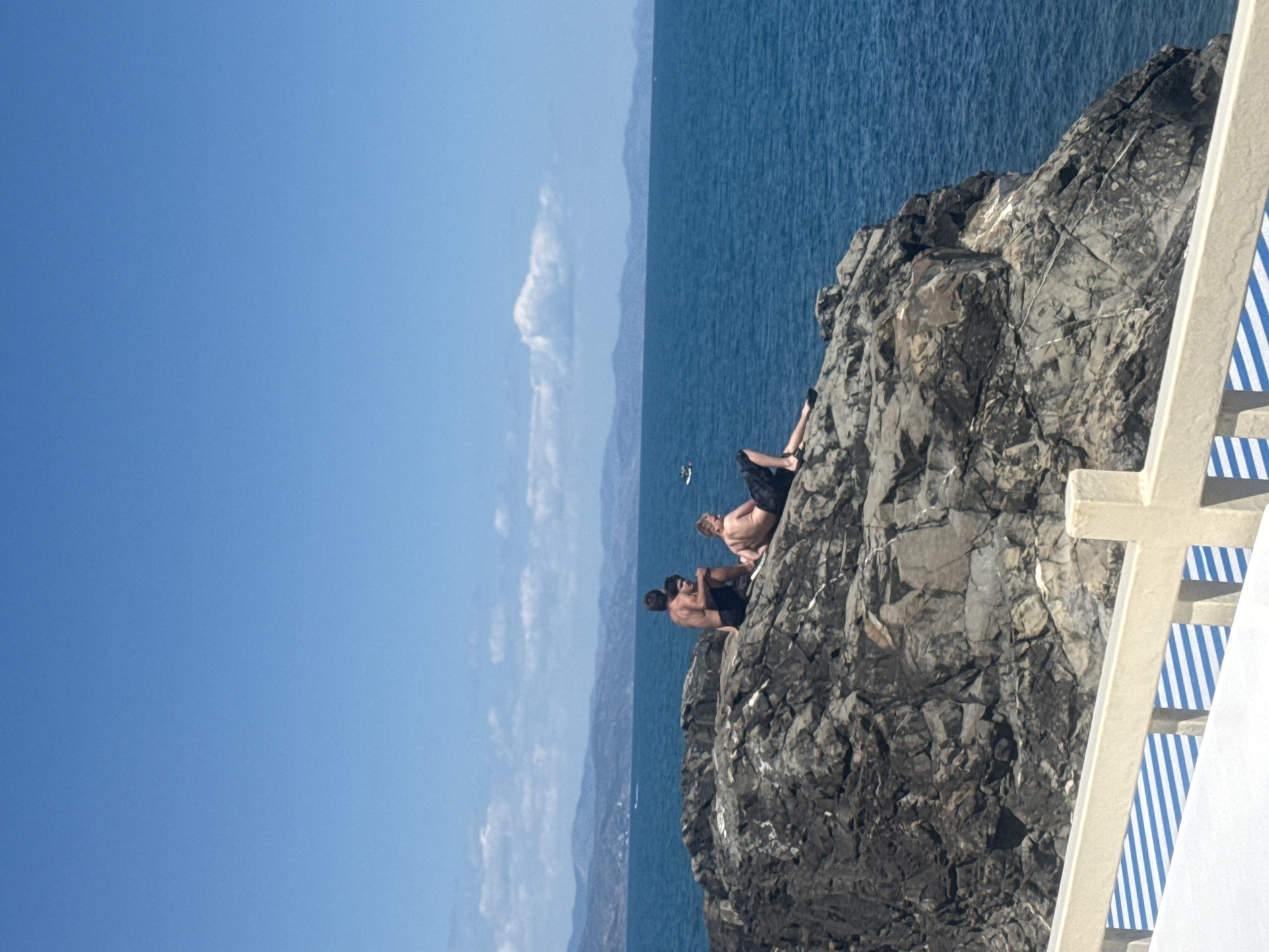 The image shows a rocky coastline with two young men sitting on the rocks overlooking the ocean. The sky is clear and blue with some white clouds in the distance. The ocean is a deep blue color. In the background, there are mountains and some boats on the water. In the foreground, there is a white railing with blue stripes. The men are both wearing swim trunks. One man is sitting with his legs crossed, and the other is lying down.