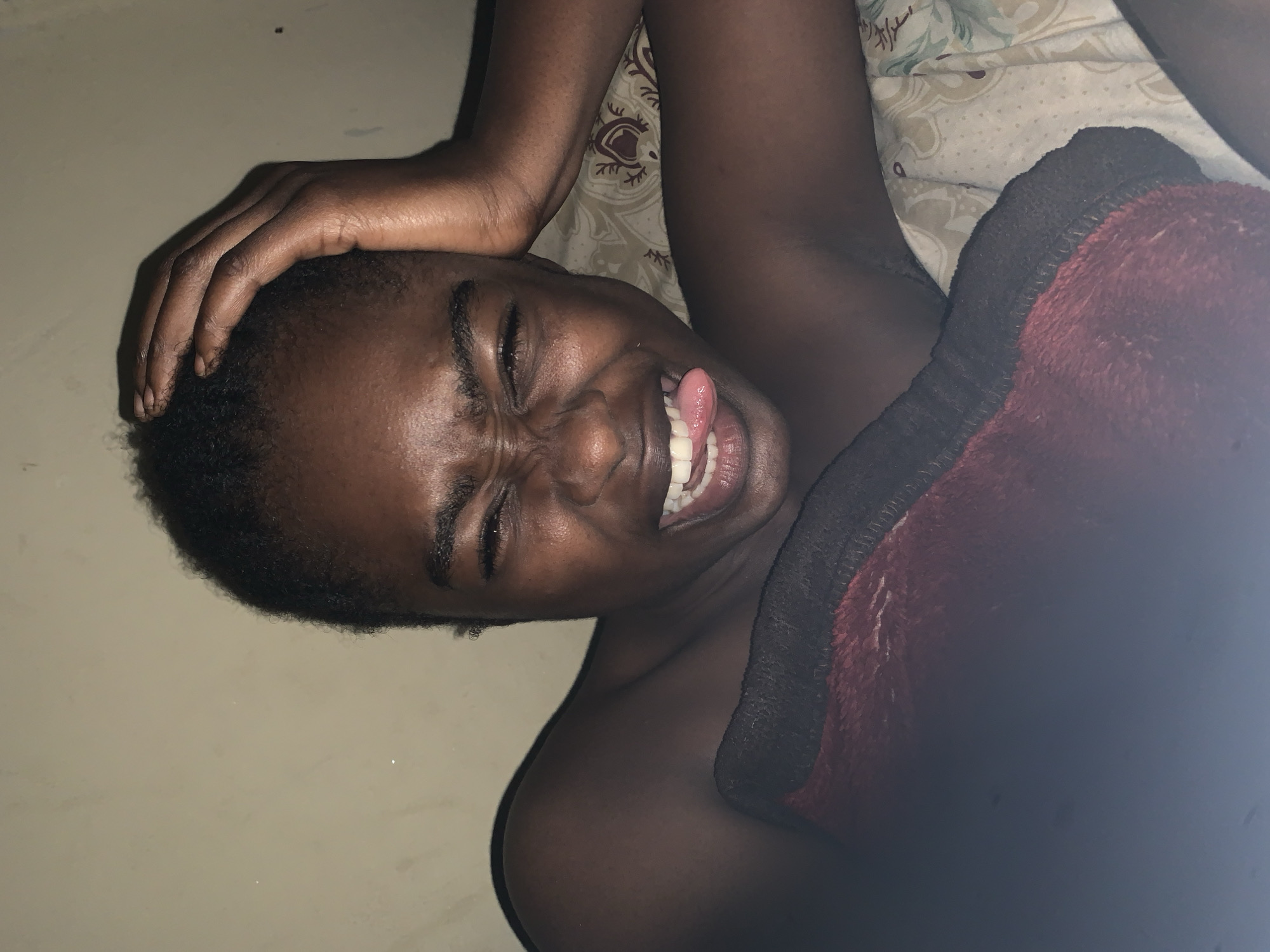 The image shows a dark-skinned individual lying on a bed with a patterned sheet and a burgundy blanket with a black border. The person is positioned on their side, facing the camera. One hand is placed on their head, while the other side of their body is partially covered by the blanket. The individual is squinting their eyes and sticking their tongue out. The background includes a plain wall. There's no way to ascertain the image's location in Rafin Par, Nigeria, from the provided information.