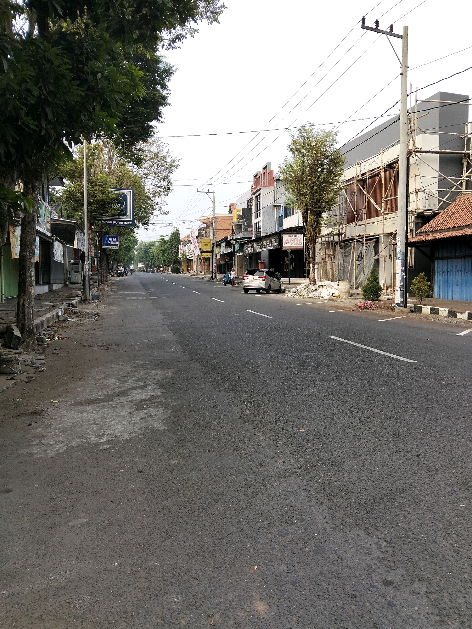 The image depicts a street scene in Purworejo, Indonesia, characterized by a paved road with a white dashed line marking the lanes, lined with buildings and trees. On the left side of the road, several trees with dense foliage partially obscure the facades of commercial establishments. One building has a sign that reads "ATM BCA". Further down the street, more trees and buildings are visible, some of which appear to be under construction with scaffolding. A silver car is parked on the right side of the road, near a building with a blue shuttered entrance. Several motorcycles are parked on the side of the road in the distance. Two individuals are walking on the sidewalk on the right side of the road, and a person is pushing a cart. The sky is overcast. The street appears relatively empty, suggesting a quiet time of day.