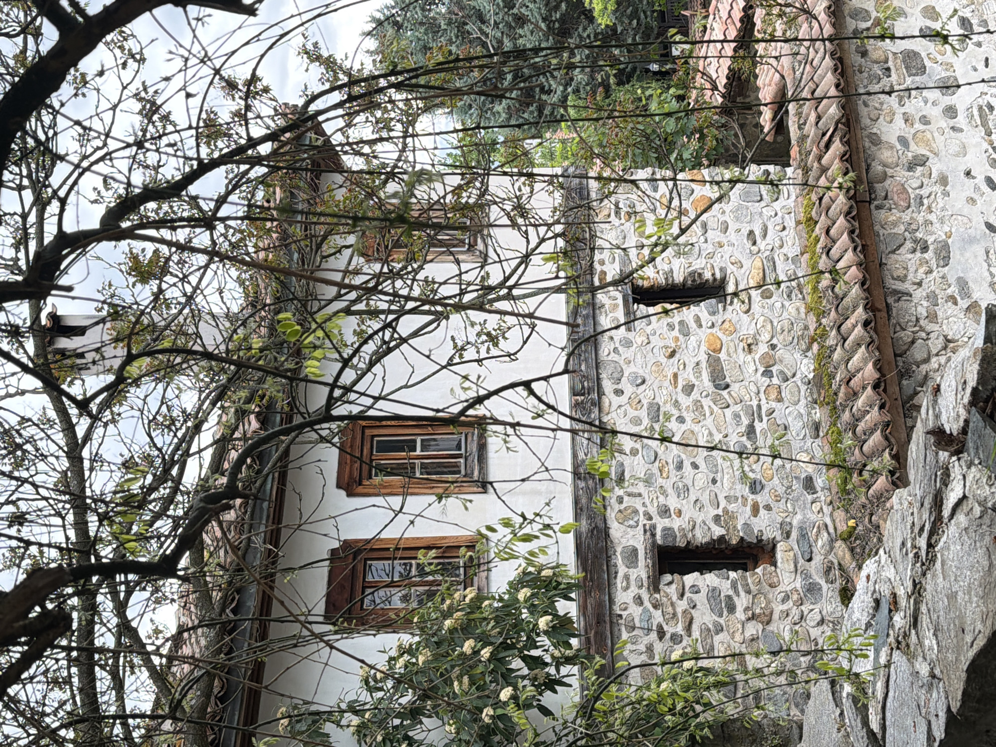 The image depicts a building in Blagoevgrad, Bulgaria. The building has both white stucco and exposed stone facade sections. The stucco section is a multi-story structure with wooden-framed windows. The stone section shows a wall constructed of rounded stones set in mortar, with narrow, rectangular openings. A roof with clay tiles is visible atop the stone section. The scene is partially obscured by bare tree branches and foliage.