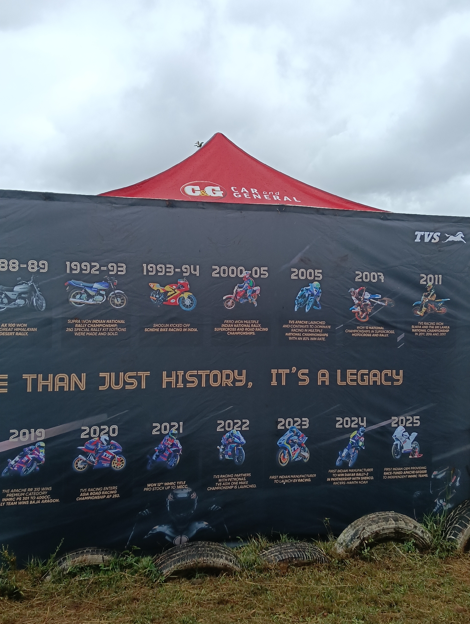 The image shows a large black banner with a timeline of TVS motorcycle racing history. The banner is set up outdoors, partially under a red tent with the "CAR and GENERAL" logo. The timeline spans from 1988-89 to 2025, with each entry featuring a motorcycle image and a brief description of achievements in racing. The banner's tagline reads, "MORE THAN JUST HISTORY, IT'S A LEGACY." In the foreground, there are tires and grassy vegetation, suggesting an outdoor event or racing venue. The image does not provide enough information to determine that it is in the city of Nairobi, Kenya.