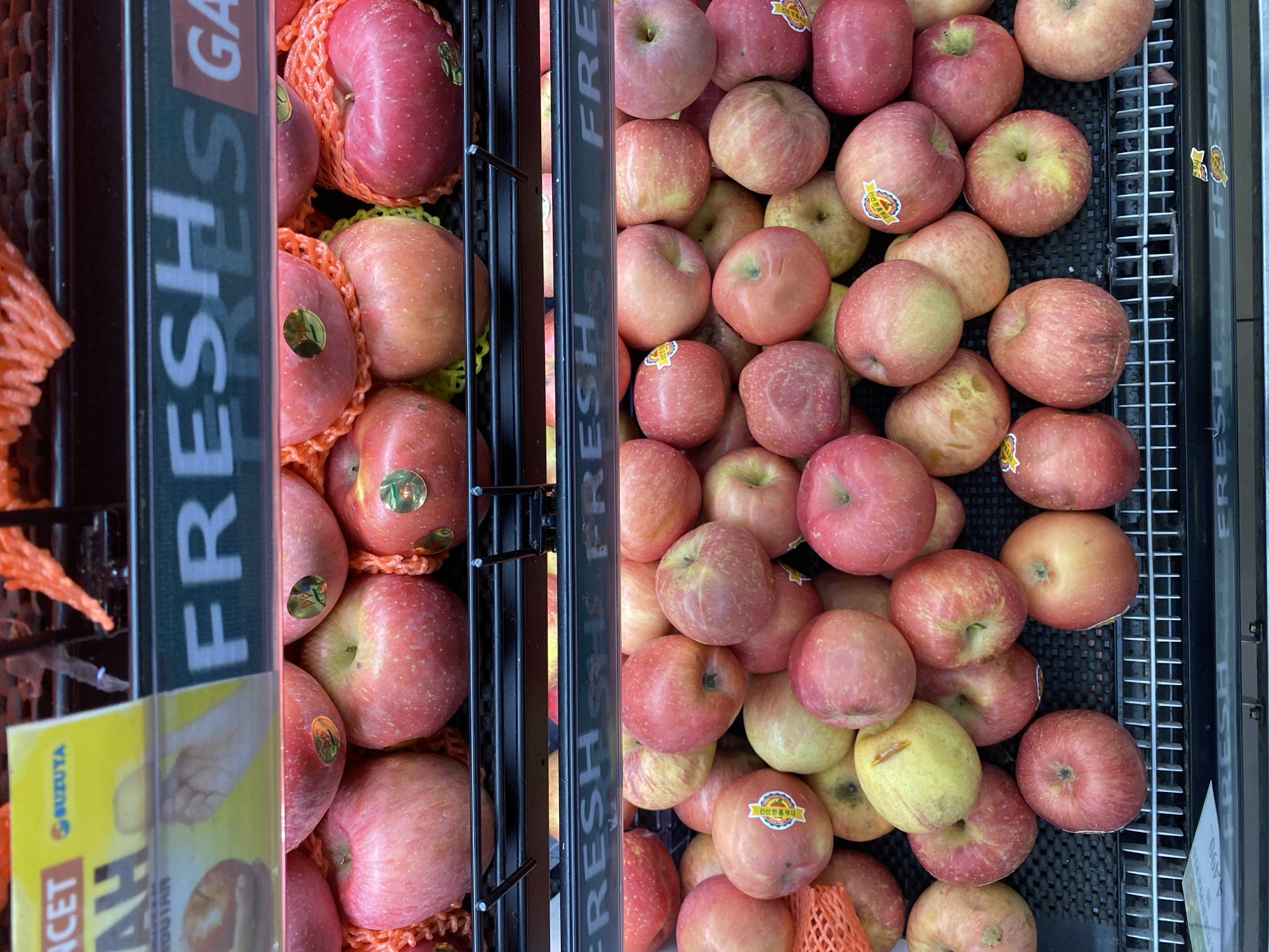 The image shows a display of apples in a market. The apples are red and pink, and they are arranged in rows on shelves. Some of the apples are wrapped in orange netting. There is a sign in the background that says "FRESH". The image appears to be taken in a grocery store or market. The text on the yellow sign in the background, which is partially visible, reads "SUZUYA". Suzuya is a department store chain in Indonesia, with branches in several cities including Banda Aceh. Therefore, the image is likely taken at a Suzuya department store in Banda Aceh, Indonesia. The overall environment appears to be indoors, with artificial lighting illuminating the produce.