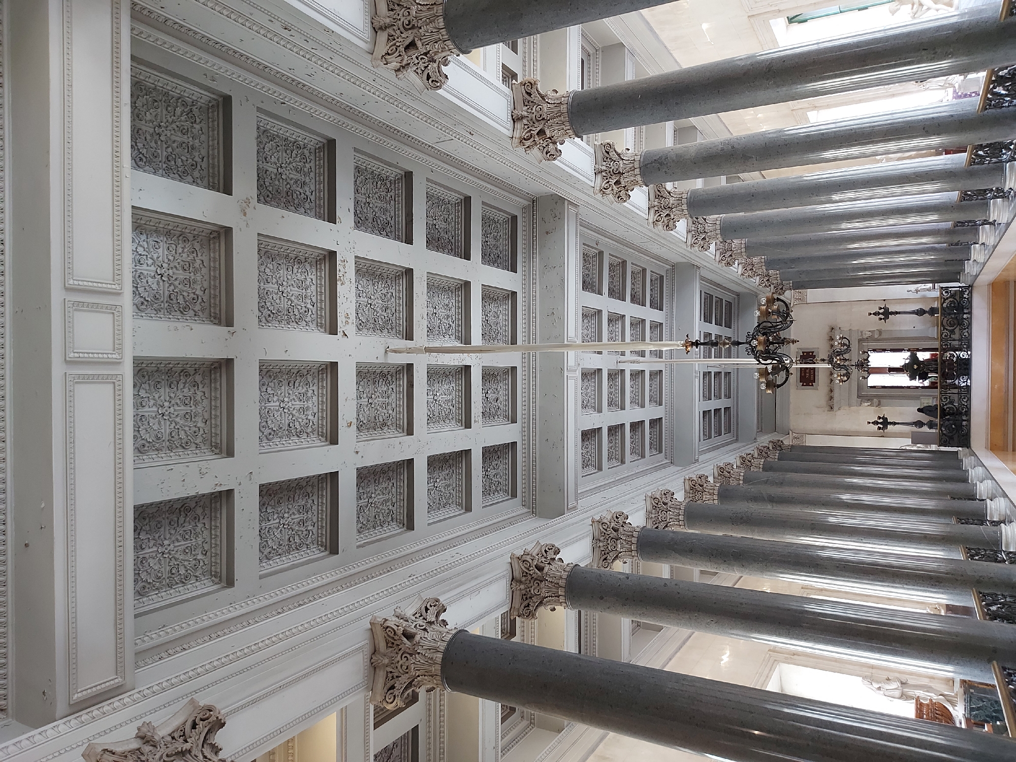 The image shows a perspective view of an interior space in Saint Petersburg, Russia. The scene is dominated by a row of gray marble columns with ornate white Corinthian capitals, running along the right side of the frame. On the left is a white ceiling decorated with square panels featuring intricate floral designs and bordered by molding. A chandelier is suspended from the ceiling, closer to the end of the corridor. In the far background, light streams in, illuminating sculptures, railings, and a marble floor, suggesting the end of the corridor opens into a larger hall.