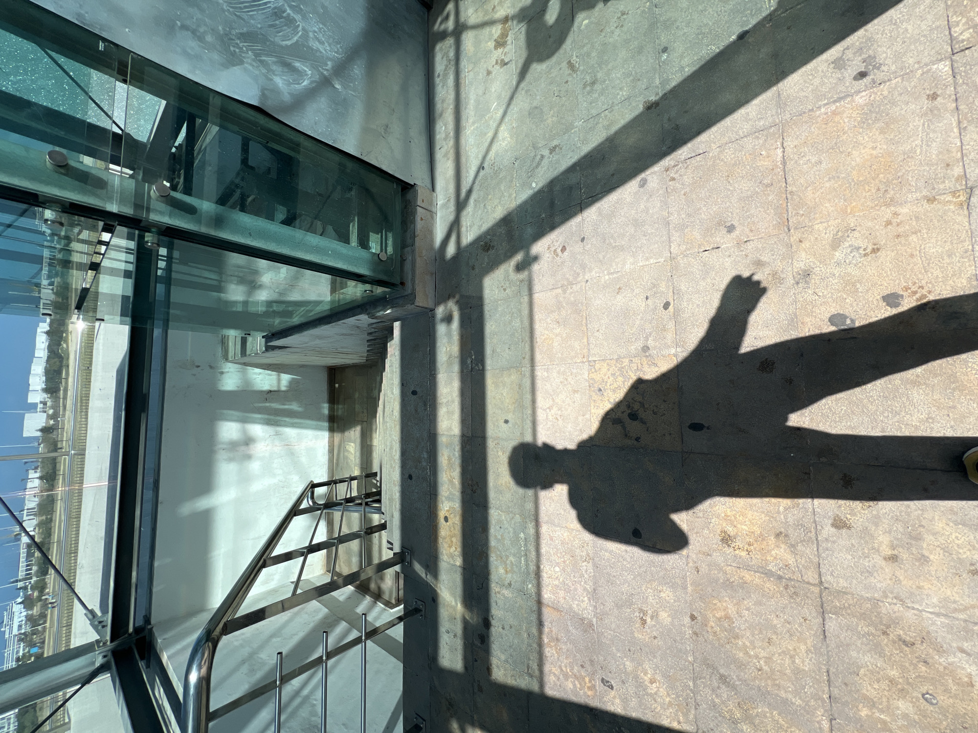 The image depicts a modern urban pedestrian access point in the city of Tanger, Morocco, specifically in a developed area such as the Corniche or near Tanger City Center, characterized by contemporary architecture and infrastructure. The scene is illuminated by strong sunlight, casting sharp shadows.
In the foreground, a tiled ground surface, light brown and grey with visible wear and dirt stains, is prominently featured. A large, dark shadow of a person, presumably the photographer, is cast across these tiles, with a small portion of a light-colored shoe visible at the bottom edge. Rectangular shadows from an overhead structure also intersect the ground.
The midground shows a concrete stairwell descending downwards. A stainless steel handrail with multiple horizontal bars runs along the left side of the stairs. The walls adjacent to the stairwell are painted an off-white or light cream, displaying some signs of age and possible water discoloration near the base. To the right, a large structure composed of tinted glass panels, supported by dark metal beams and fasteners, encloses the stairwell entrance. This glass reflects ambient light and partially obscures elements beyond.
Through the glass structure and above the stairwell, an exterior urban environment is visible. A multi-lane road or highway, featuring a white concrete barrier, runs horizontally across the background. Beyond the road, several large, modern buildings with light-colored facades are discernible under a clear sky. Some sparse green landscaping is also present between the road and the distant buildings. No other individuals are visible within the frame. The overall impression is a functional, modern urban transit or access point under bright daylight conditions.