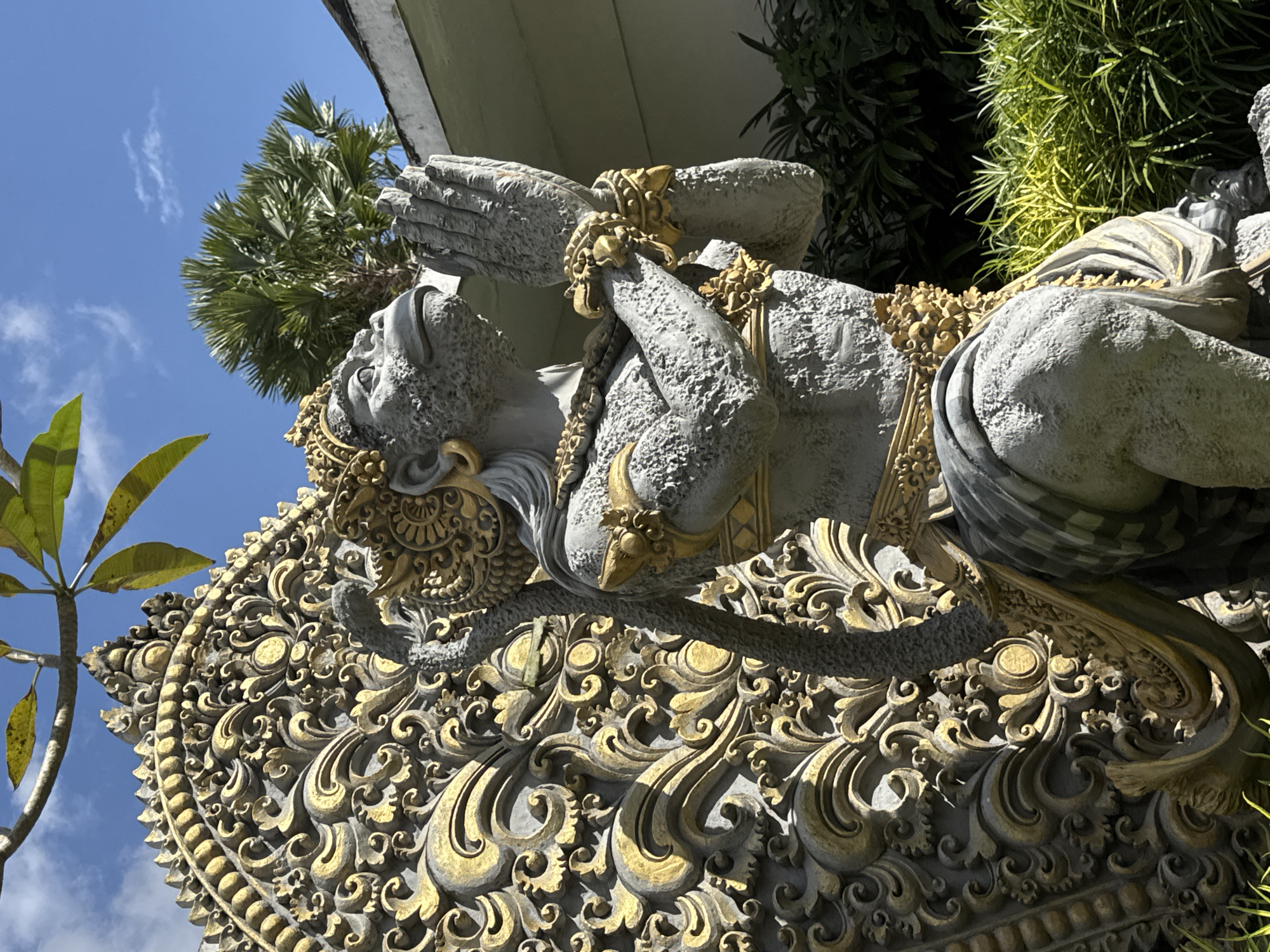 A stone statue of a monkey-like figure with praying hands is positioned in front of an ornate, golden-colored relief carving. The statue is gray and textured, adorned with gold accents on its headdress, jewelry, and clothing. It is kneeling with its hands pressed together in front of its chest, looking upwards. Behind the statue, a large, intricate carving features swirling, organic patterns in gold and gray. The scene is outdoors, with lush green foliage and palm trees visible in the background, under a clear blue sky with scattered white clouds. The location is identified as Tegallalang, Indonesia.
