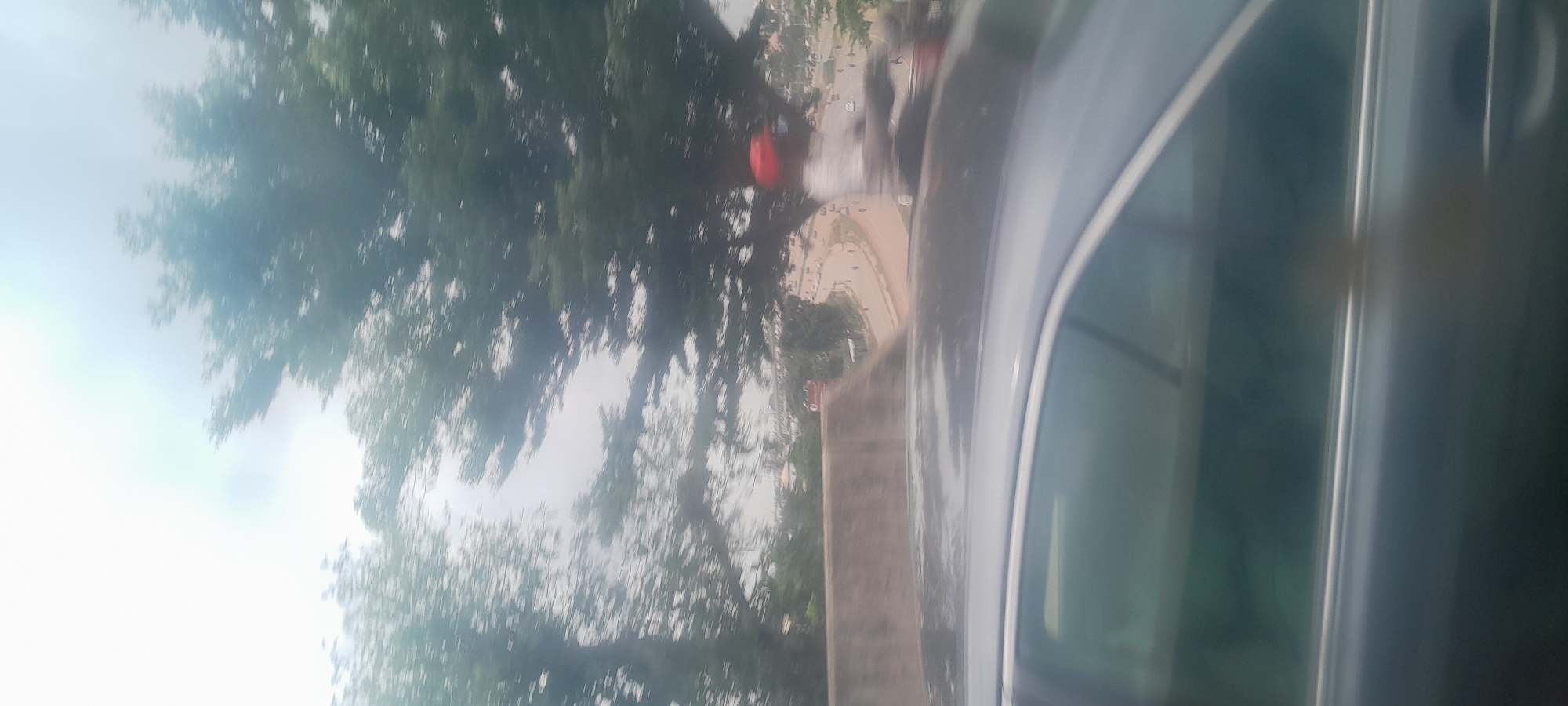 The image captures a street scene in Kugbo, Nigeria, with a focus on a gray car in the foreground, seen from the side. Beyond the car, a road curves to the right, with several motorcycles and a few cars visible. The road is lined with trees, some of which are quite large and leafy, obscuring parts of the sky. There are buildings in the background, some appearing to be single-story structures with red roofs. A person wearing a red cap and a white shirt is visible standing on the side of the road, appearing to be engaged in some activity. The overall impression is one of a bustling street in a city with significant greenery.