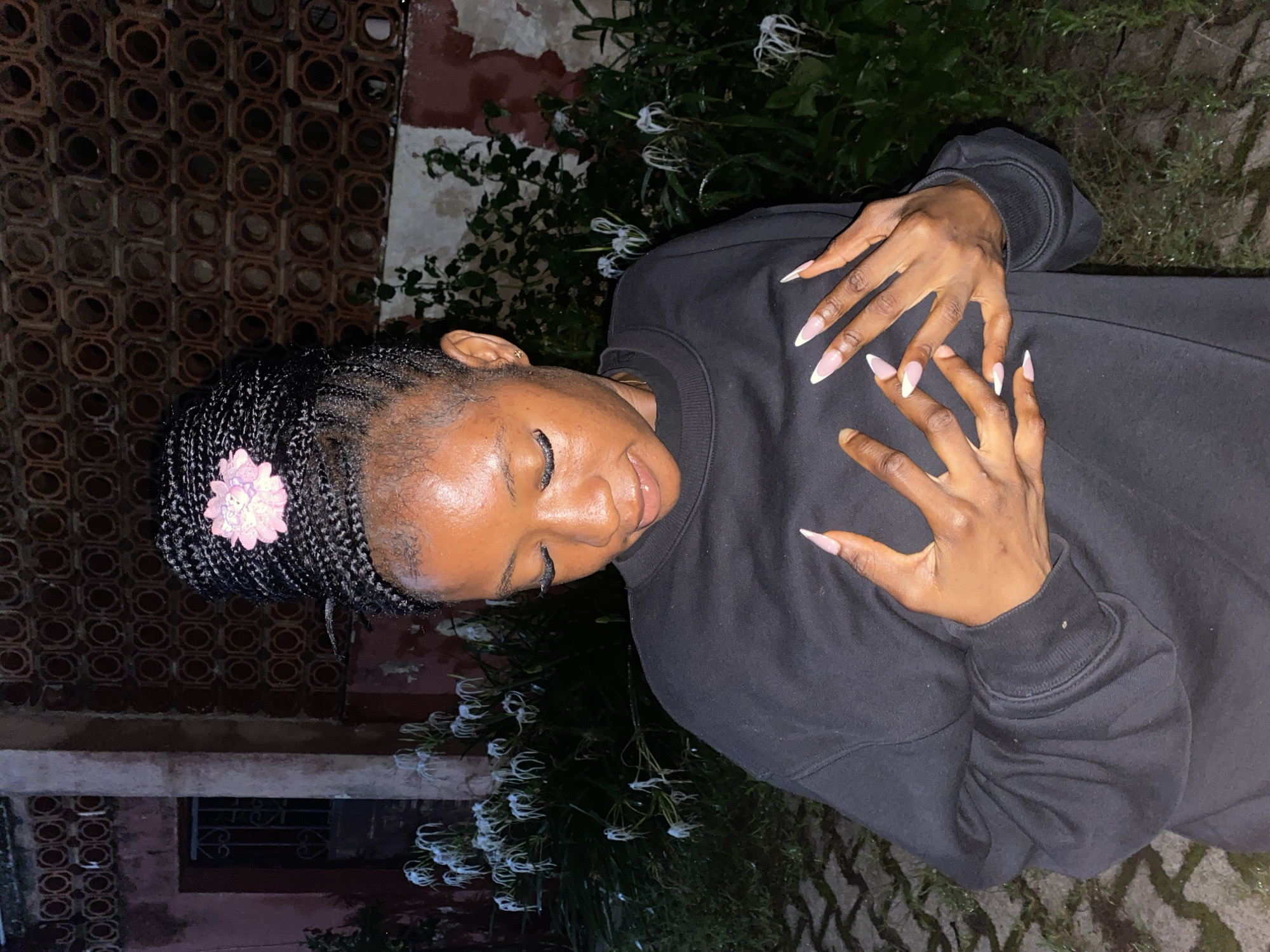 The image depicts a young Black woman with her hair styled in braids and a pink flower accessory. She is wearing a black sweatshirt and has long, light-colored acrylic nails. Her hands are positioned over her chest. The background features a building with patterned brickwork, lush green foliage, and white flowers. The ground appears to be paved with interlocking stones. While the image captures a specific moment, there are no explicit indicators to pinpoint its location within the city of Jos, Nigeria.