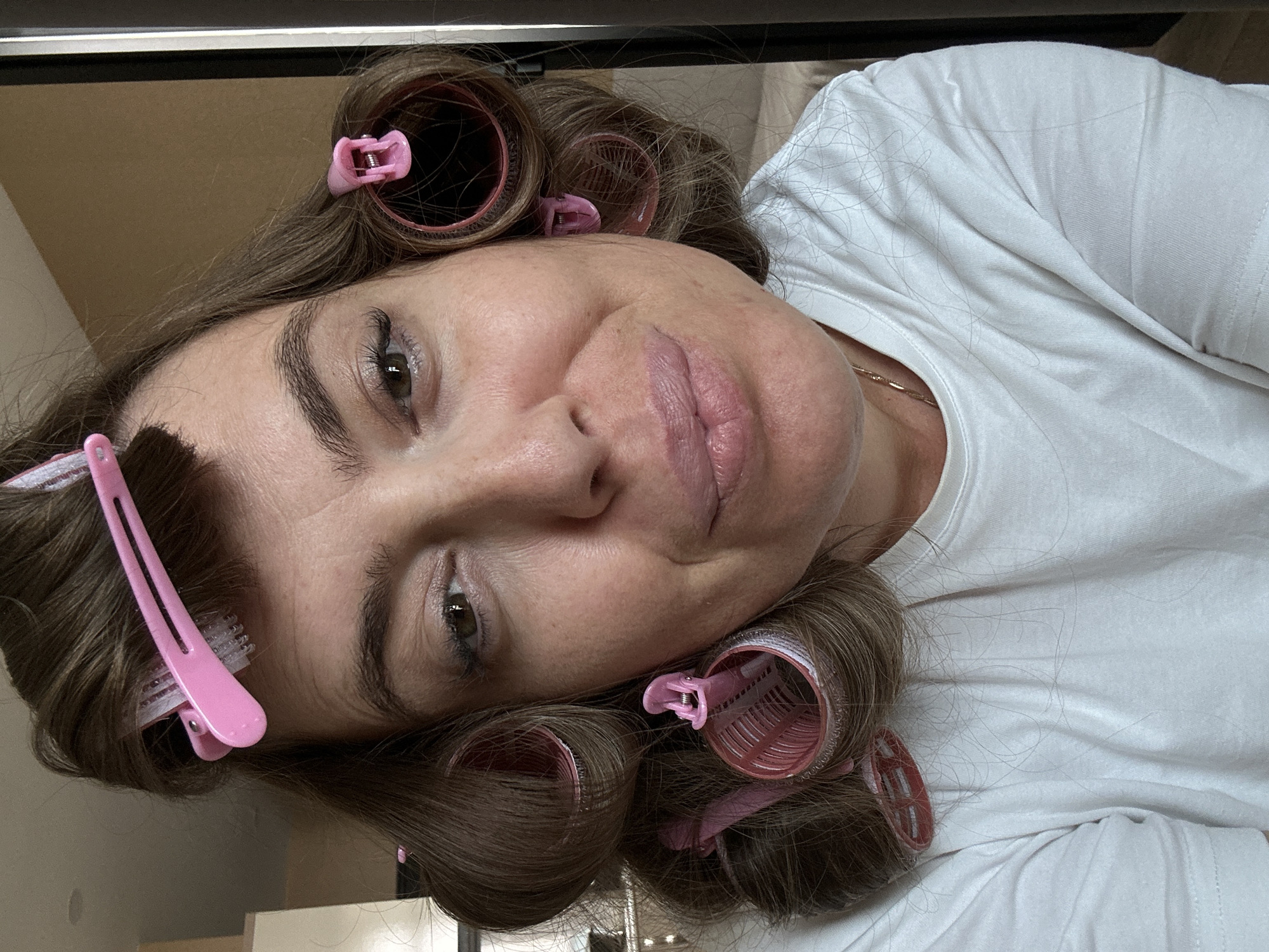 A woman with brown hair styled in rollers and pink hair clips is shown in a close-up, front-facing shot. She is wearing a white t-shirt and a gold necklace. The background appears to be an indoor setting, possibly a bedroom, with a neutral-colored wall and a glimpse of furniture. There is no specific identifiable landmark or feature to indicate the location is Batumi, Georgia.