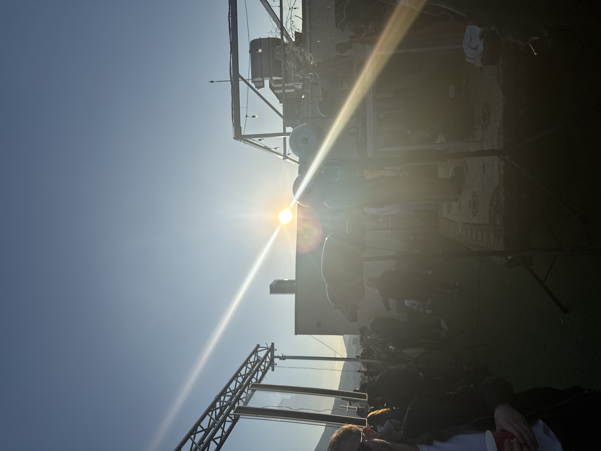 The image depicts an outdoor event, likely a concert or festival, taking place under a clear, sunny sky in Sofia, Bulgaria. The scene is illuminated by the intense, low-angle light of the sun, which is prominently positioned in the upper-middle left of the frame, casting strong lens flares and bright sunbeams across the image.
A large crowd of people fills the lower-right and mid-right portions of the frame, some sitting and others standing on what appears to be green artificial turf. In the immediate foreground, to the bottom right, part of a person's head and shoulder are visible, holding a red cup.
Event infrastructure is prominent, with a large metal truss structure extending from the lower-left towards the mid-left. Near the center, to the right of the sun, several large, rounded objects resembling stage speakers or lighting fixtures are visible. In the distance, behind these structures and partially silhouetted, is a tall, rectangular building, indicating an urban setting. Various poles, stands, and wires associated with event production are also scattered throughout the venue.
The strong direct sunlight creates high contrast, with significant areas of overexposure and deep shadows that obscure finer details within the crowd and background structures. The time of day appears to be late afternoon or early evening, given the sun's position and the dramatic lighting. Text on the event equipment is largely obscured by the glare and shadows, making it illegible.