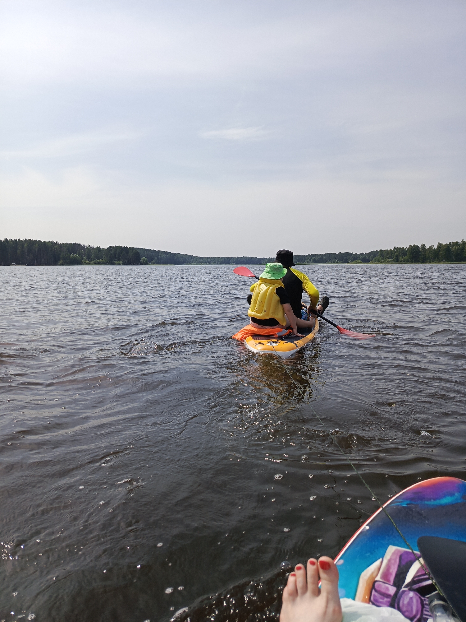Two individuals are on a stand-up paddleboard on a body of water. The person in the front is wearing a yellow life vest and a green hat. The person behind them is wearing a black and yellow long-sleeved shirt and a black hat. Both are facing away from the camera, with the person behind holding a red paddle. In the foreground, a person's foot with red nail polish is visible resting on another board. The background consists of a treeline and a hazy sky. The scene appears to be a recreational activity on a lake or river, possibly in Chaykovsky, Russia, given the context of the city.