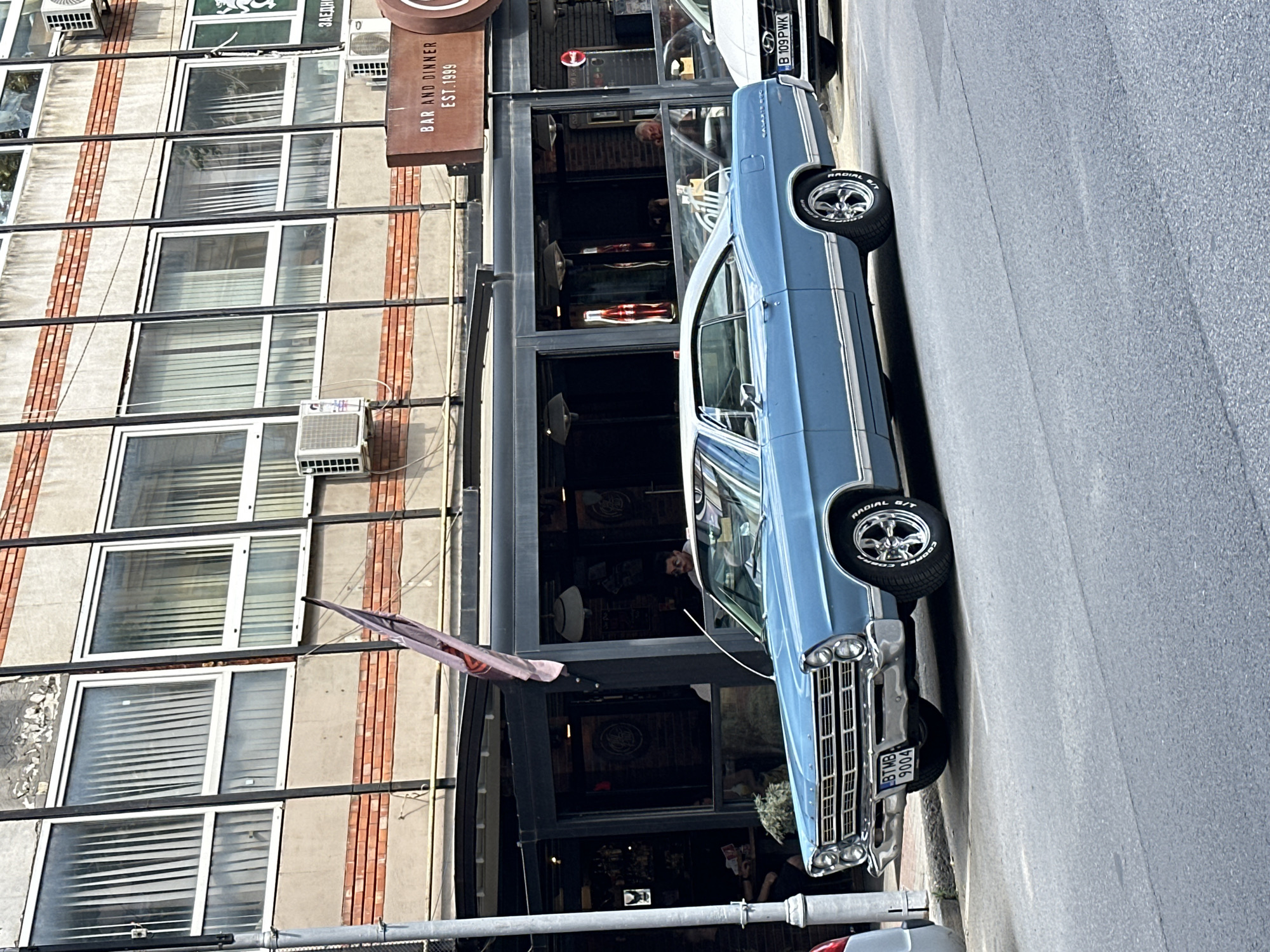 The image captures a classic light blue Ford Galaxie 500 parked on a city street, likely in Veliko Tarnovo, Bulgaria, given the license plate "BTMB 9004" which resembles Bulgarian plates and "B 109PWK" which is a Bulgarian license plate. The car is positioned on the right side of the road, angled slightly towards the viewer. In the background, a multi-story building with a concrete facade and red brick accents stands. The building features several large windows with vertical blinds. Below the building, a street-level establishment with a dark facade, large glass windows, and outdoor seating is visible. A sign above this establishment reads "BAR AND DINNER EST. 1999". Several people are seated inside or near the establishment, including a man with glasses visible through the car window. Another car, a white modern sedan, is parked to the right of the classic car, with its license plate clearly visible. The street surface is asphalt, showing some wear and markings. The overall scene suggests a typical urban environment on a bright day.