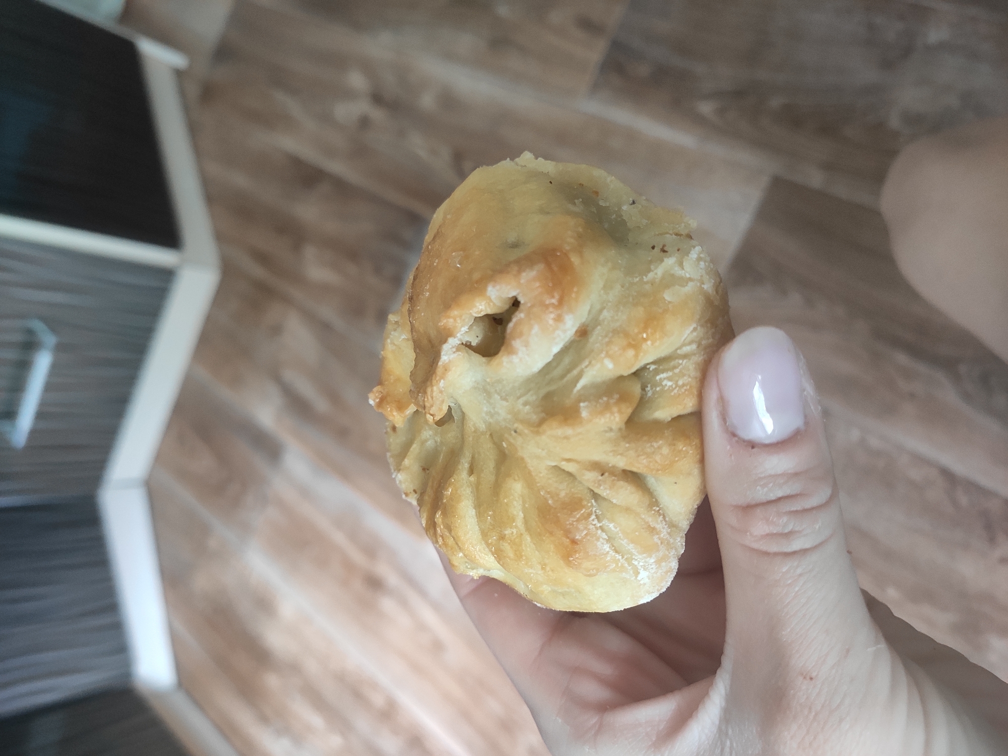 A hand holds a baked bun/pastry. The location is plausibly Sterlitamak, Russia, as the AI model does not have enough information to confirm this with certainty, but the image lacks any geographic or identifying data to substantiate this claim. The pastry appears to be a light golden brown color and has a somewhat twisted or folded appearance. The background consists of a wood-look floor and part of a white and gray cabinet.