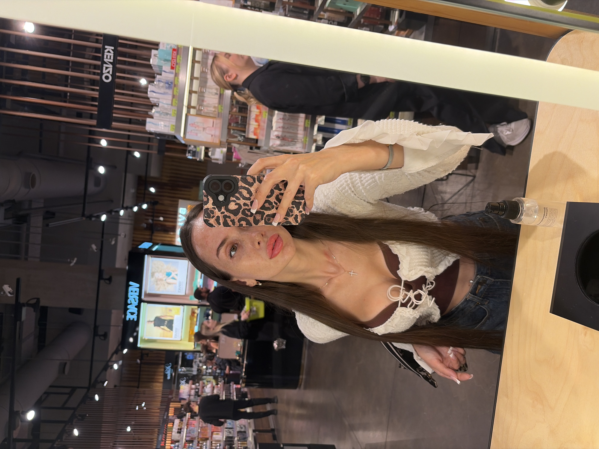 The image is a selfie taken by a young woman in a brightly lit store. She is holding a smartphone with a leopard-print case. She has long, dark brown hair and is wearing a white ruffled top over a brown corset-like garment, and blue jeans. A delicate silver necklace with a cross pendant is visible. Her makeup is natural, emphasizing her eyes and lips.
In the background, reflected in a large mirror, a woman in a black jacket and jeans is visible, as well as shelves stocked with various products, suggesting a retail environment like a beauty or fashion store. The store appears to be indoors. There are also displays with images of models, and overhead lighting. The overall atmosphere seems casual and contemporary.
Visible text in the image includes "KENZO" on a black sign hanging from the ceiling, and the word "ABPVVCF" (likely a mirrored or distorted brand name) on a display panel. The setting appears to be a commercial space in Saint Petersburg, Russia, given the provided location context. The lighting suggests it is daytime or the store is well-illuminated. The woman's expression is neutral, and the image quality is clear.