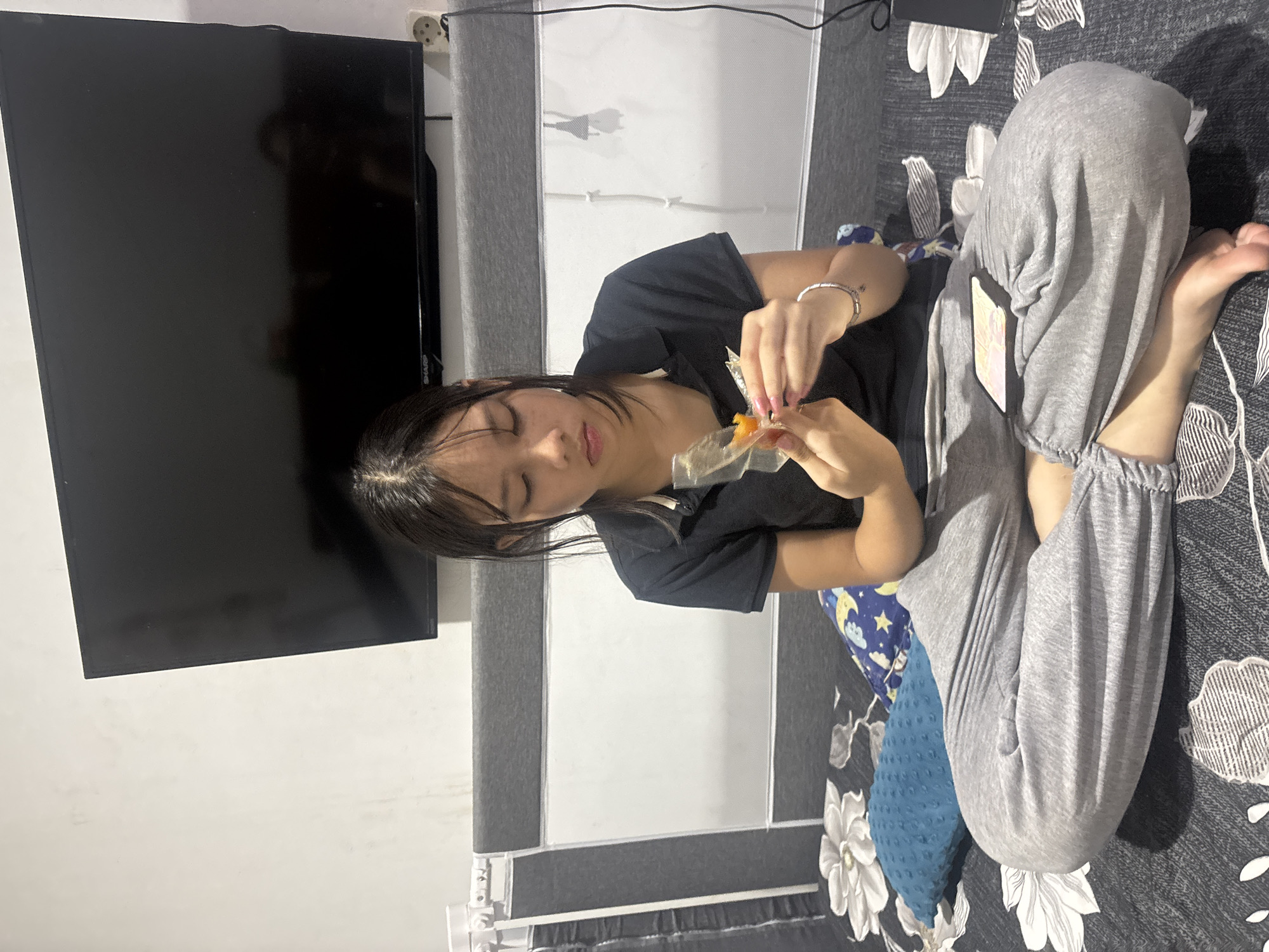 An Indonesian female teenager, wearing a black polo shirt and gray sweatpants, sits cross-legged on a bed with a dark patterned comforter and a blue textured pillow. She is holding a small orange fruit, possibly a physalis, in a clear plastic wrapper, and appears to be examining or peeling it. Her gaze is directed downwards. Behind her, a large flat-screen television is mounted on a white wall above a grey headboard. The overall environment suggests an indoor setting, likely a bedroom, in the city of West Jakarta, Indonesia.