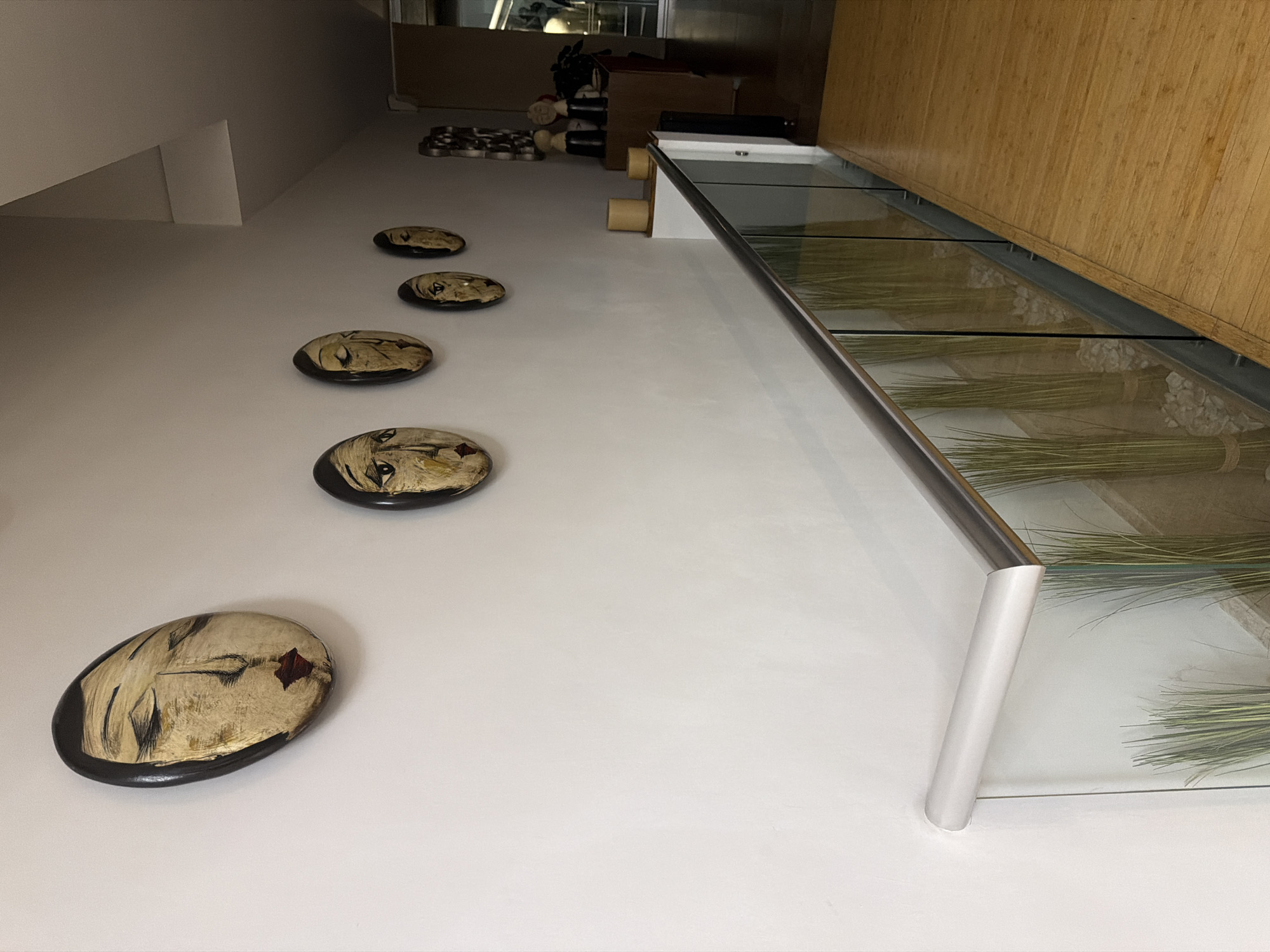 The image shows an interior space in San Bernardino Tlaxcalancingo, Mexico, featuring decorative wall art. Five oval-shaped art pieces with face depictions are mounted on a white wall. Above the art, a glass-topped display case with metallic supports runs along the top of the wall. The case contains arrangements of grass. The wall is adjacent to a bamboo-paneled wall. In the background, there is furniture and decor items, including what appears to be chess pieces.