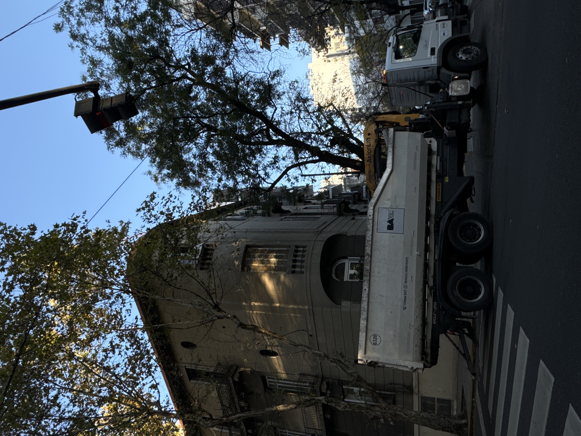 A medium-angle street-level view shows a white dump truck parked at a crosswalk along a street in Buenos Aires, Argentina. The truck's bed displays the "BA" logo, signifying it belongs to the city's government. A yellow hydraulic arm is attached to the truck. The crosswalk marks the intersection of a side street with a main thoroughfare. The building on the left side of the image is a gray, multi-story, pre-war building, partially obscured by trees and branches. A traffic light is visible on the left, hanging over the intersection. The sky is clear and blue.