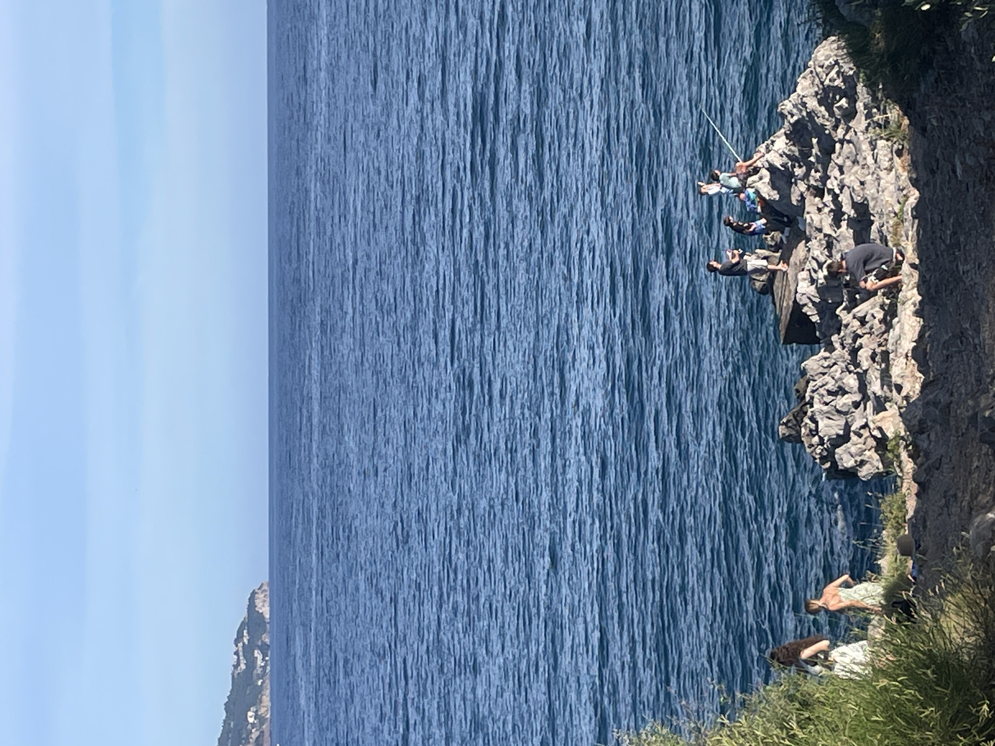 The image depicts a coastal scene with a group of people fishing on a rocky outcrop in the Adriatic Sea. The water is a deep blue, with gentle waves. In the distance, a lush green coastline with buildings is visible. The foreground shows a rocky terrain with some vegetation. Several individuals are present: on the rocky outcrop, a group of at least five people are seen fishing with rods, and another person is sitting on the rocks. Closer to the viewer, on the left side, two women are standing near the edge of the water. One woman is wearing a light-colored dress, and the other is in a green patterned dress. A dark-colored hat is placed on the ground near them. The lighting suggests it is a sunny day. The location is identified as being near Budva, Montenegro, likely on the coast overlooking the Adriatic Sea.