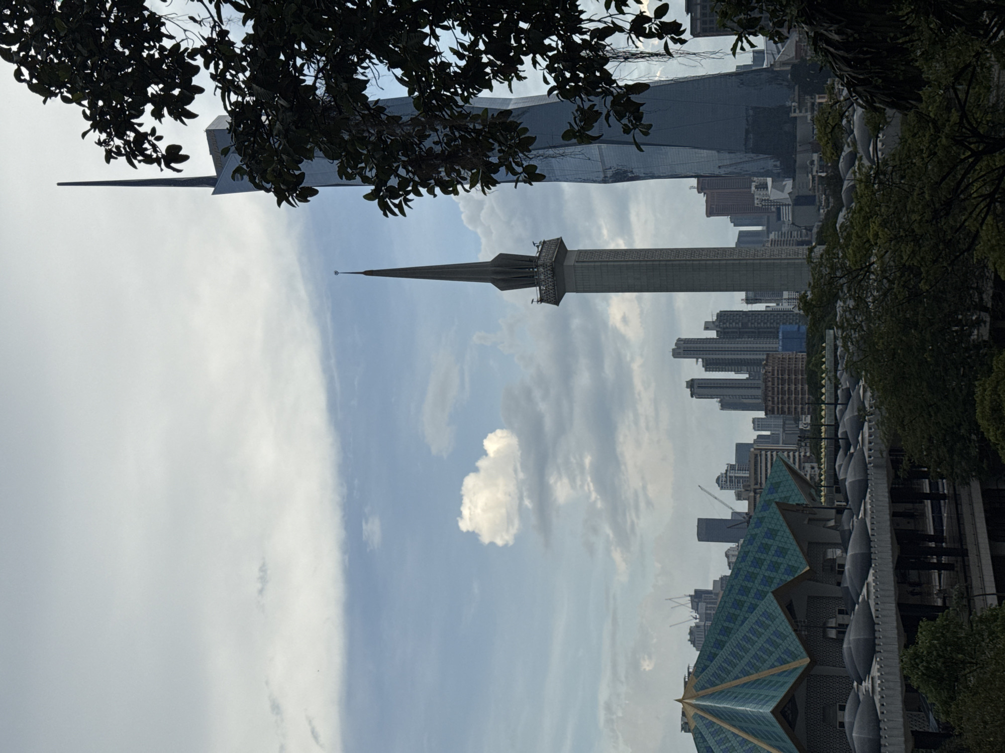 The image is a low-angle shot of Kuala Lumpur, Malaysia, showcasing notable structures amidst a partly cloudy sky. The Islamic Arts Museum Malaysia is in the foreground, recognizable by its distinctive green and blue tiled roof and row of bulbous domes. A tall minaret, part of the mosque, rises prominently. Behind the mosque, a dense cluster of skyscrapers forms the city skyline, including the Petronas Twin Towers, partially obscured by trees. The overall environment suggests an urban setting with architectural landmarks.