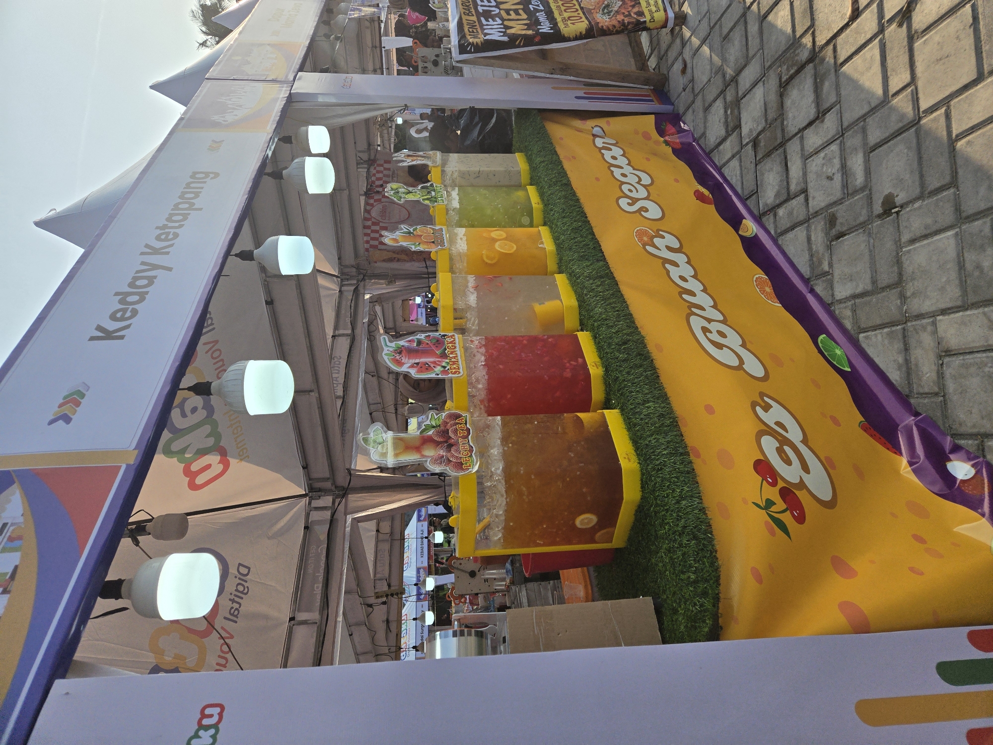 The image shows an outdoor food stall in Sungailiat, Indonesia, likely at a festival or market. The stall is decorated with a large yellow banner featuring the words "Es Buah Segar" (Fresh Fruit Ice) and images of fruits like cherries and strawberries. In front of the banner are several large, clear dispensers filled with colorful beverages, each labeled with fruit names such as "Lechy Tea," "Semangka" (Watermelon), and drinks with orange and lime. The dispensers are placed on a green artificial grass mat. Above the stall, a white tent provides shade, with the name "Keday Ketapang" visible on a sign. Several white LED lights hang from the tent structure. In the background, people are visible, and there is a sign board displaying a menu, with the text "MENU BARU MIE JE... MEN..." partially visible. The ground is paved with interlocking concrete tiles. The lighting suggests it is late afternoon or early evening, with the sun casting shadows.