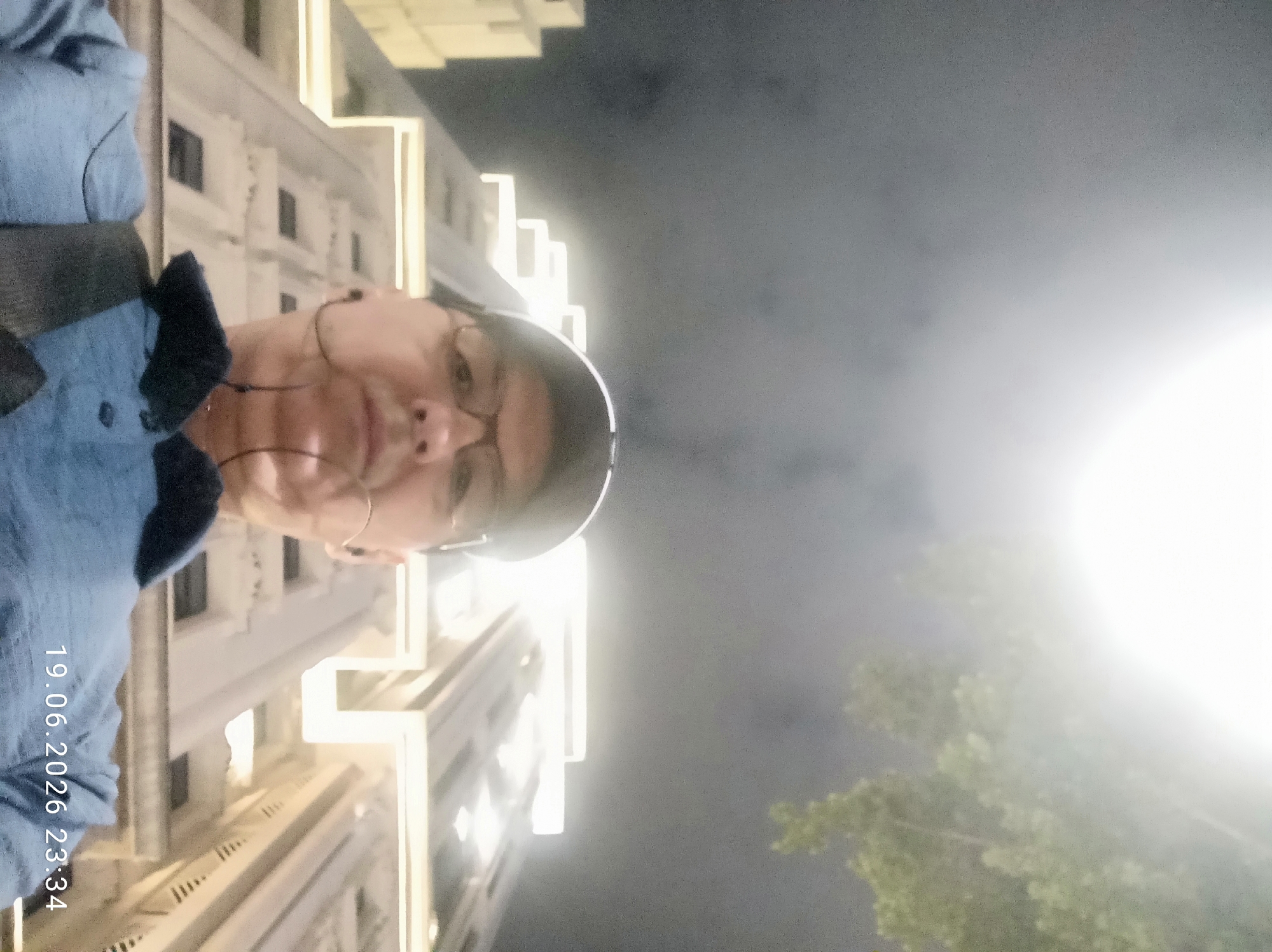 A man is taking a selfie at night in front of illuminated buildings. He is wearing a blue shirt, glasses, and a baseball cap. He has earbuds in his ears. The timestamp "19.06.2026 23:34" is visible in the lower right corner of the image. The background shows tall buildings with lights and some tree branches at the top. The sky is dark and hazy.