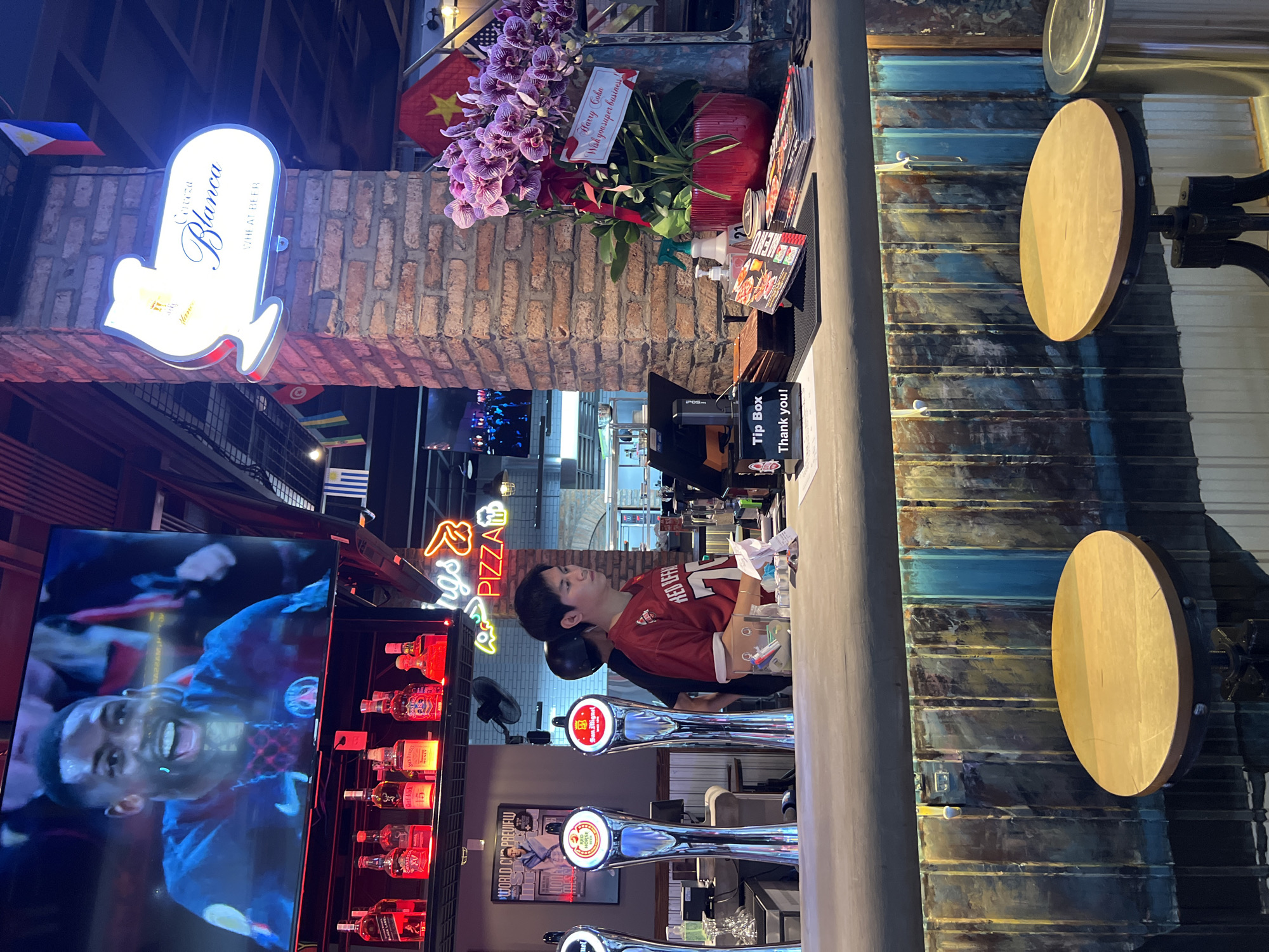 The image shows the interior of a bar, likely in Bắc Nha Trang, Vietnam, based on the Vietnamese flag present. A person wearing a red soccer jersey is visible behind the bar counter. On the back wall are numerous bottles of liquor displayed with red neon lights, a large TV showing soccer, and a neon sign that reads "Wings & Pizza." Beer taps are also visible on the counter. A sign that says "Cerveza Blanca Wheat Beer" with an arrow points towards the bottom. There are also flowers in a red vase and a sign that reads "Harry Cohn Wish you super business!" and a tip box that reads "Thank you!". Along the wall are decorative wooden circles.