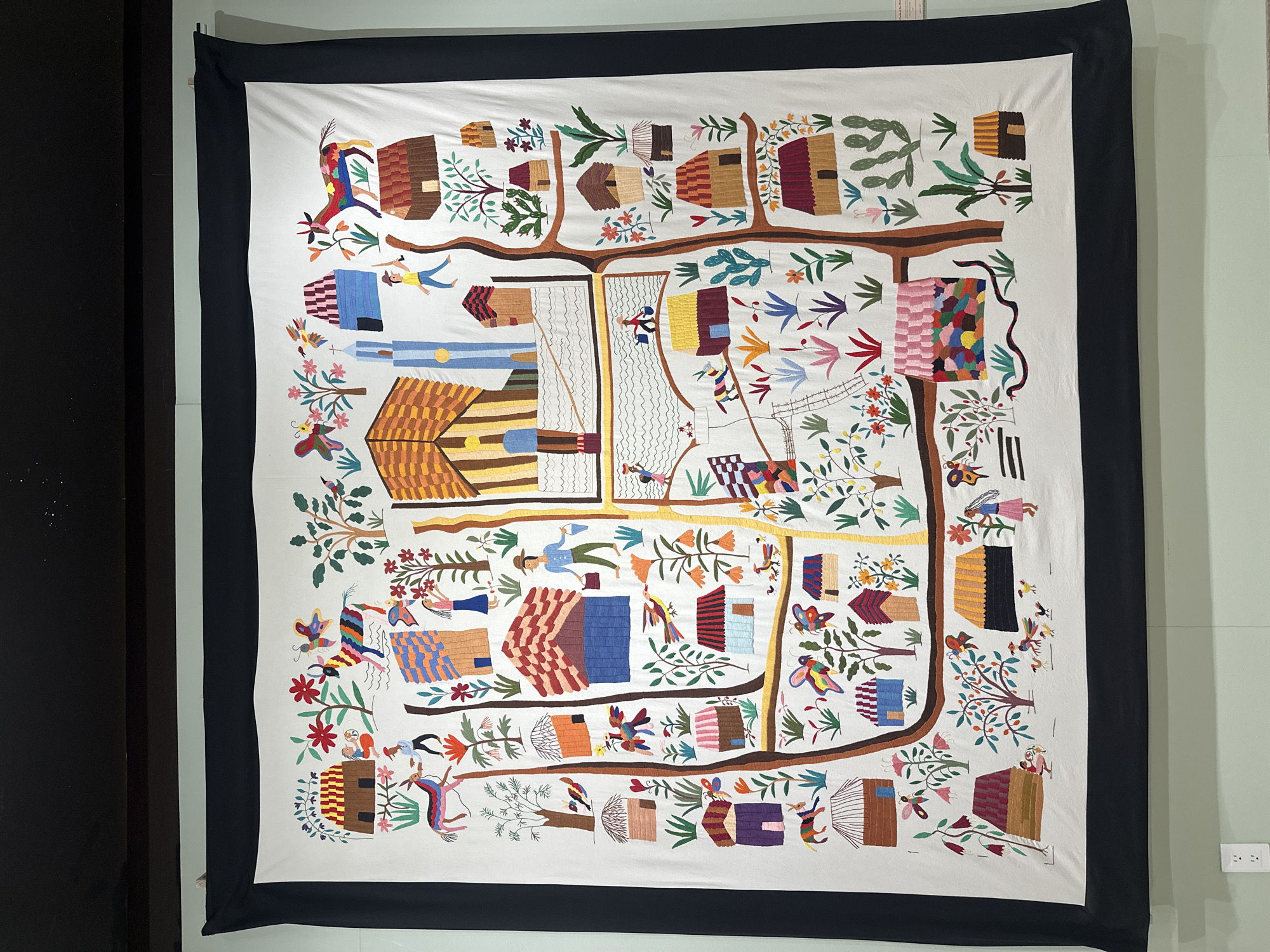 The image shows a large, rectangular textile hanging on a pale green wall. The textile has a white background with a detailed, colorful embroidered or appliqued depiction of a village scene. The scene features houses with various roof styles and colors, trees, plants, and human figures engaged in different activities. Roads or paths are visible, connecting the houses and areas. The textile has a wide, dark border. The wall has an electrical outlet. To the left of the textile is a dark, vertical surface, possibly a wall or a piece of furniture. The scene is indoors, likely in a museum or gallery. The image doesn't contain enough information to identify the location as Mexico City.