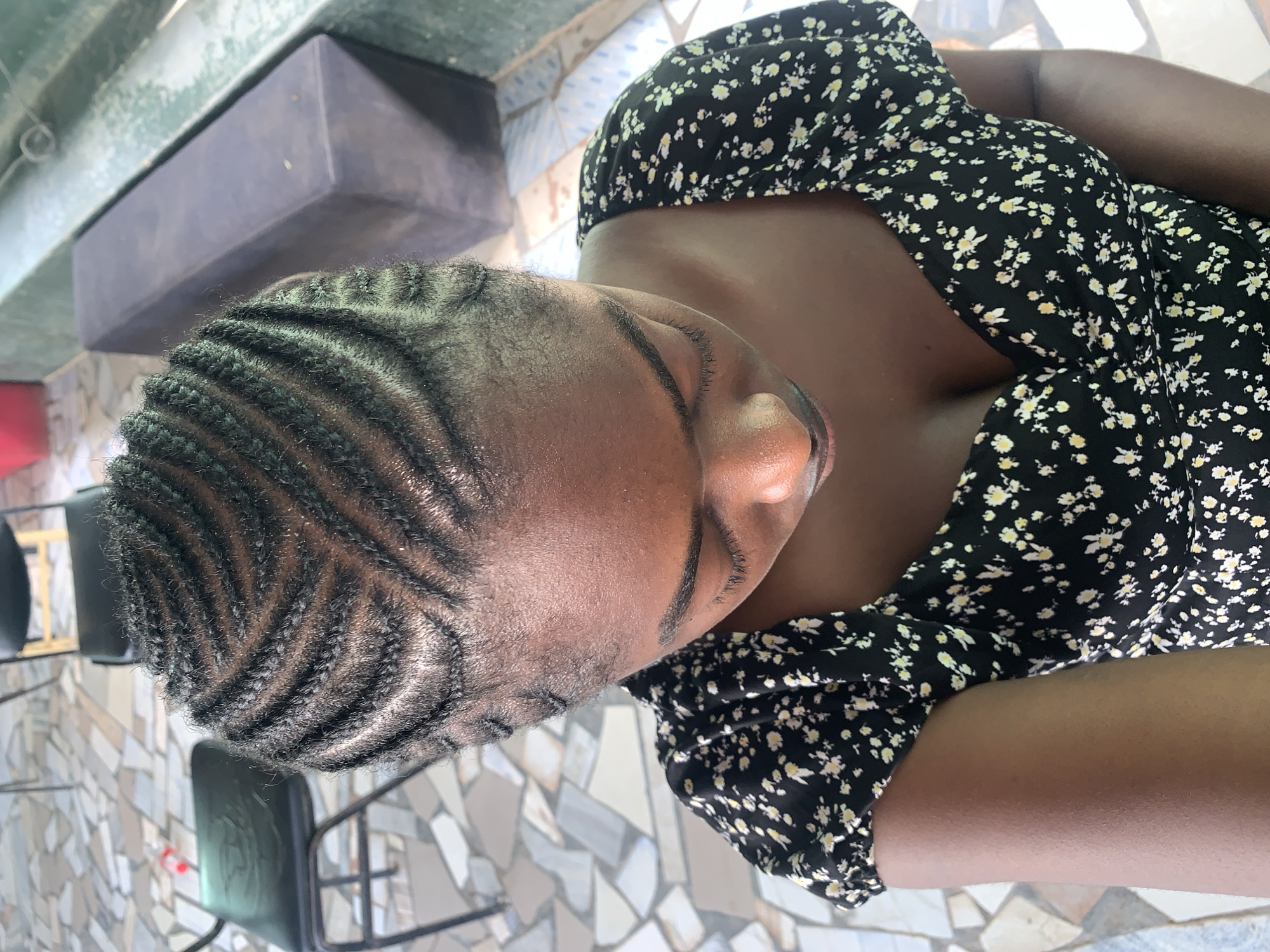 The image, rotated 90 degrees clockwise, captures a close-up view of a person from the chest up, with their head slightly bowed and turned to the right. The individual has dark skin and features meticulously braided cornrows on their scalp, arranged in a distinctive zigzag or wavy pattern. They are wearing a black dress adorned with a delicate pattern of small white and yellow flowers.
The setting appears to be an indoor space, possibly a salon or a waiting area at the New Stadium in Nigeria. The background reveals a mosaic-like tiled floor with irregular shapes in shades of white, grey, and brown. Parts of dark-colored chairs with slender metal frames are visible towards the bottom and right of the frame. Above the person's head, a dark grey, rectangular object, possibly a shelf or structural beam, is present.
The activity depicted is a still pose, likely to showcase the person's freshly done hairstyle. The lighting is bright and diffused, suggesting it is daytime. No visible text is present in the image. The overall impression is one of neatness and calm, highlighting the intricate hair design.