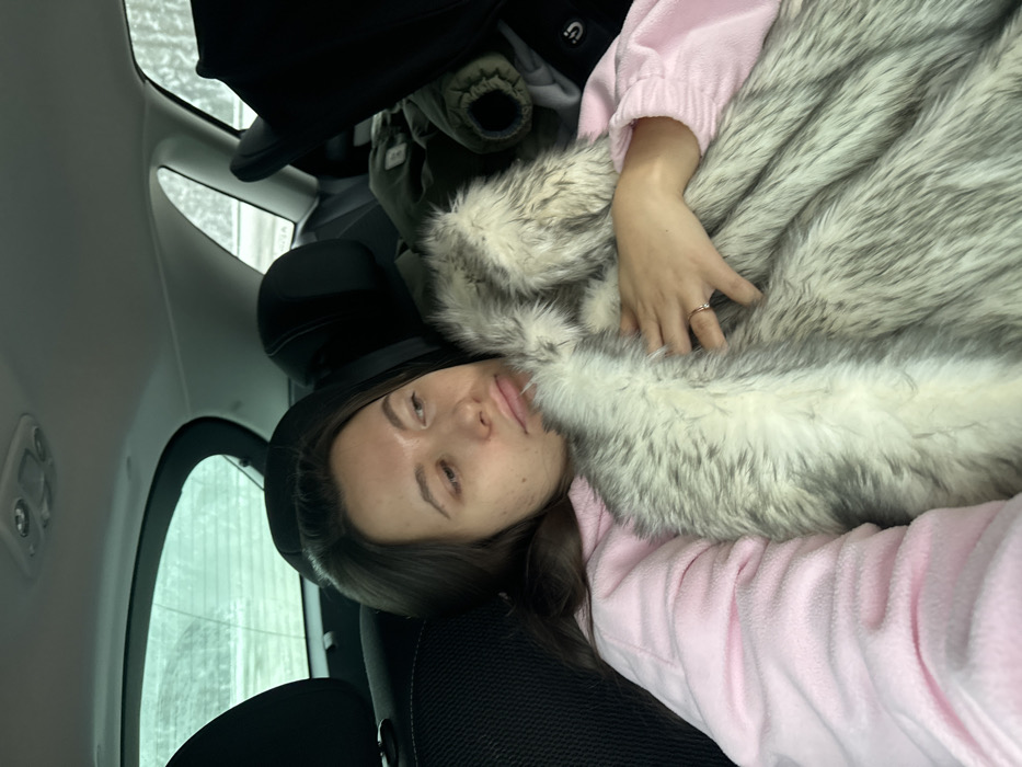 A female subject is depicted reclining in the rear passenger seat of a vehicle in Selyatino, Russia. She has long, dark brown hair and is wearing a light pink long-sleeved garment. She is holding or wrapped in a grey and white faux fur blanket, with her right hand visible on the blanket, displaying a simple gold-colored ring on her left ring finger. Her gaze is directed upwards and to the right, exhibiting a neutral to slightly pensive expression. The car interior features dark headrests on the front seats and a light-colored ceiling with a central light fixture. Through the rear windows, a blurry outdoor scene consistent with a winter landscape, possibly snow-covered, is visible. A dark green jacket and a portion of a dark car seat or similar object are present on the seat to the subject's right.