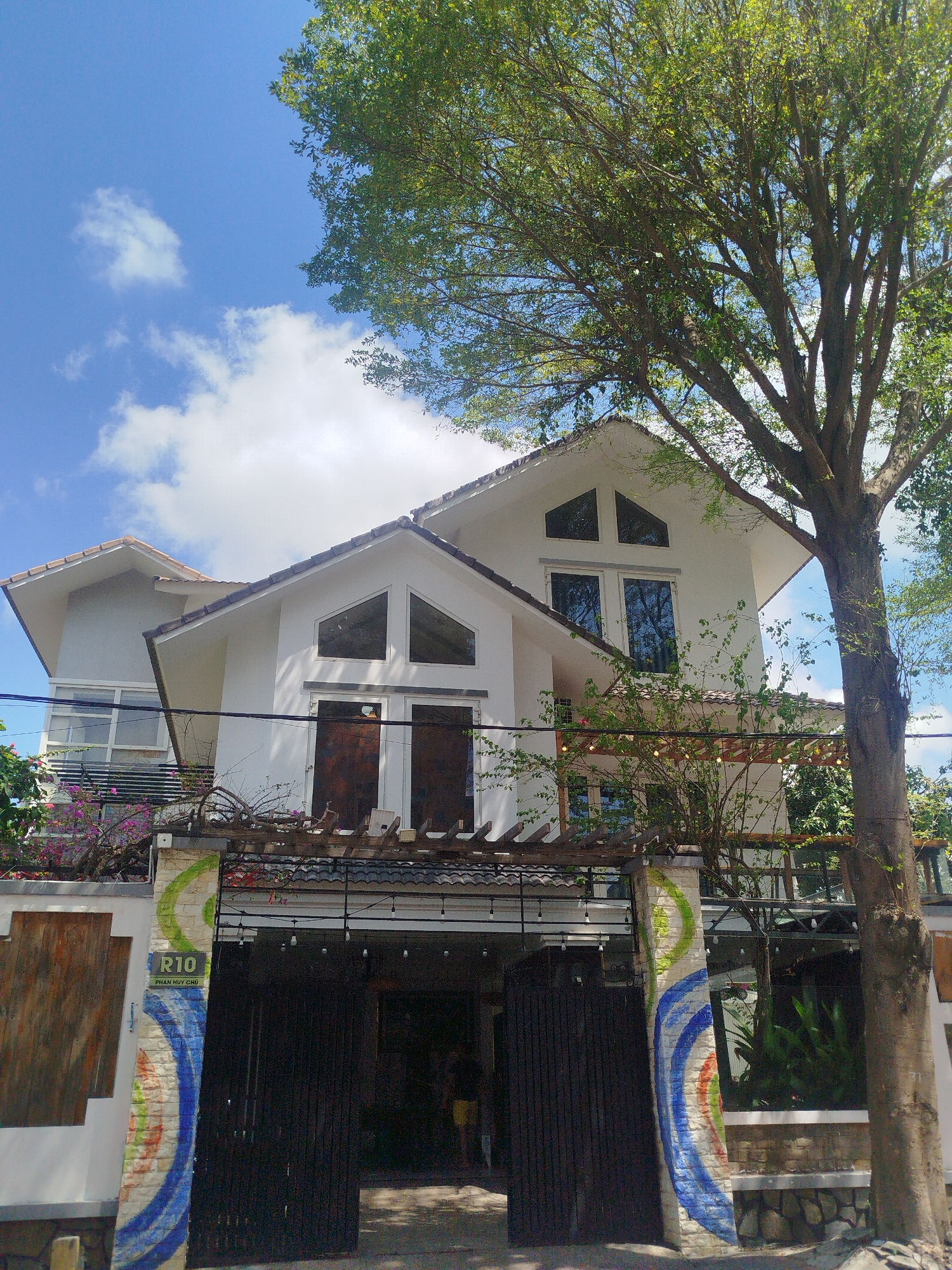 A daytime exterior shot shows a two-story white building with dark wood doors and windows, framed by a tall tree on the right and a wall with graffiti on the left. The building has a gabled roof and multiple windows. A pergola with string lights is above the entryway. A sign reads "R10 PHAN HUY CHU." The sky above is blue with scattered clouds. The building is located in Ho Chi Minh City, Vietnam. A person in yellow shorts is partially visible inside the building's entrance. The graffiti on the wall consists of concentric blue, green, and red arcs.