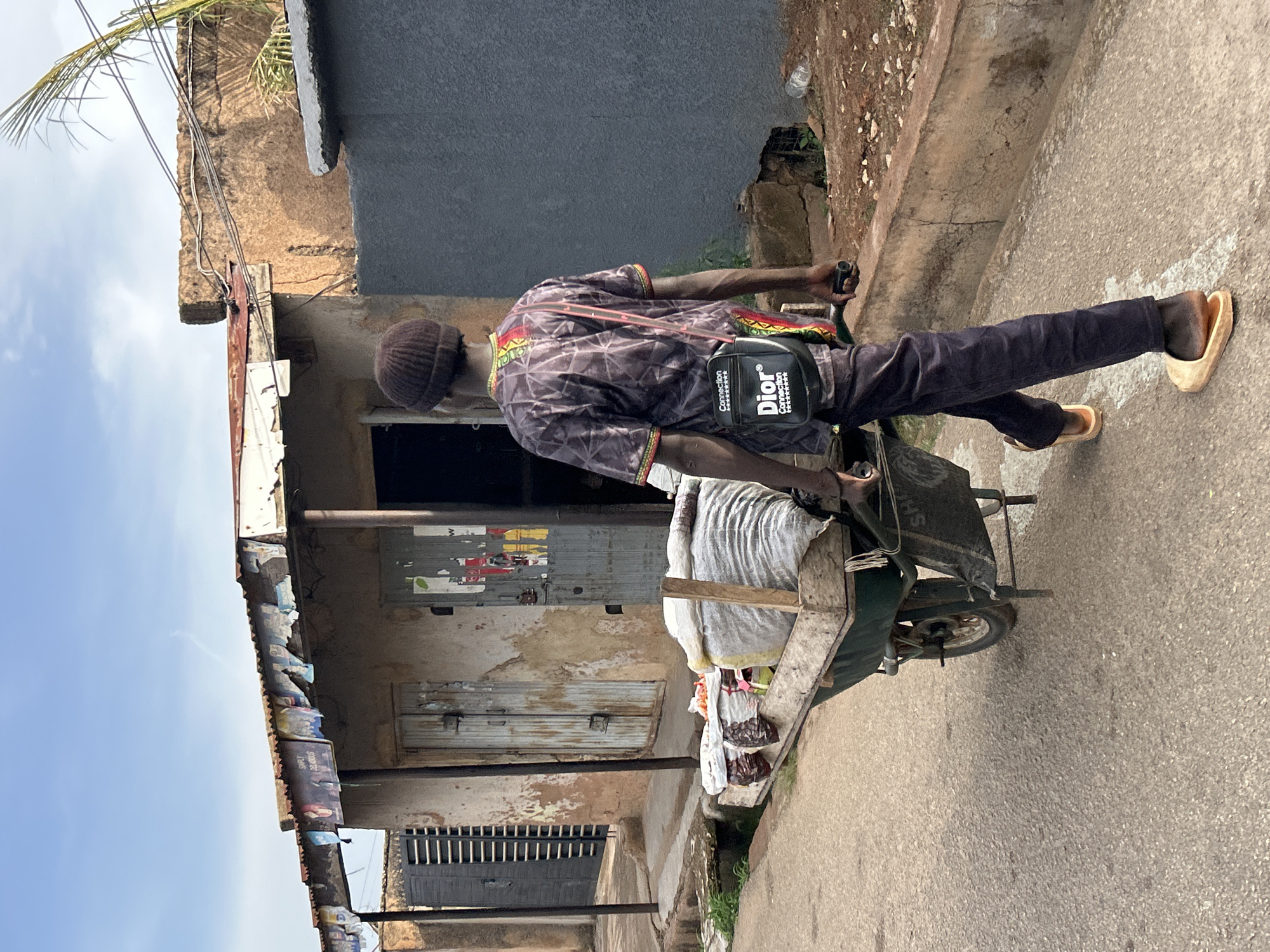 A man is depicted from a back-left perspective, pushing a loaded wheelbarrow along a paved street in Jos, Nigeria, under a bright, partly cloudy sky. He is dressed in a dark brown knit beanie, a dark, patterned long-sleeved shirt with red, yellow, and green trim on the cuffs and collar, dark trousers, and light-colored slip-on sandals or clogs. A black messenger bag is worn across his body, prominently displaying "Dior Connection" and "Connection" in white text.
The man pushes a dark green, single-wheeled handcart with wooden side supports. It carries a large, light-colored sack and several other items, possibly in plastic bags, visible at the front. The street is paved with a light grey, aggregate surface, flanked by a concrete curb.
To the left of the street are several old, weathered buildings. Their facades show signs of age, with peeling paint, exposed concrete or stucco, and multiple doors, some metal and some wooden, featuring visible locks. Posters and advertisements cling to the walls and a corrugated metal roof, which is also partially covered with old packaging. Overhead, electrical or communication wires crisscross the scene, connecting to the structures. A large green palm frond is visible near the upper left corner, suggesting a tropical environment. The clear daylight indicates it is daytime.