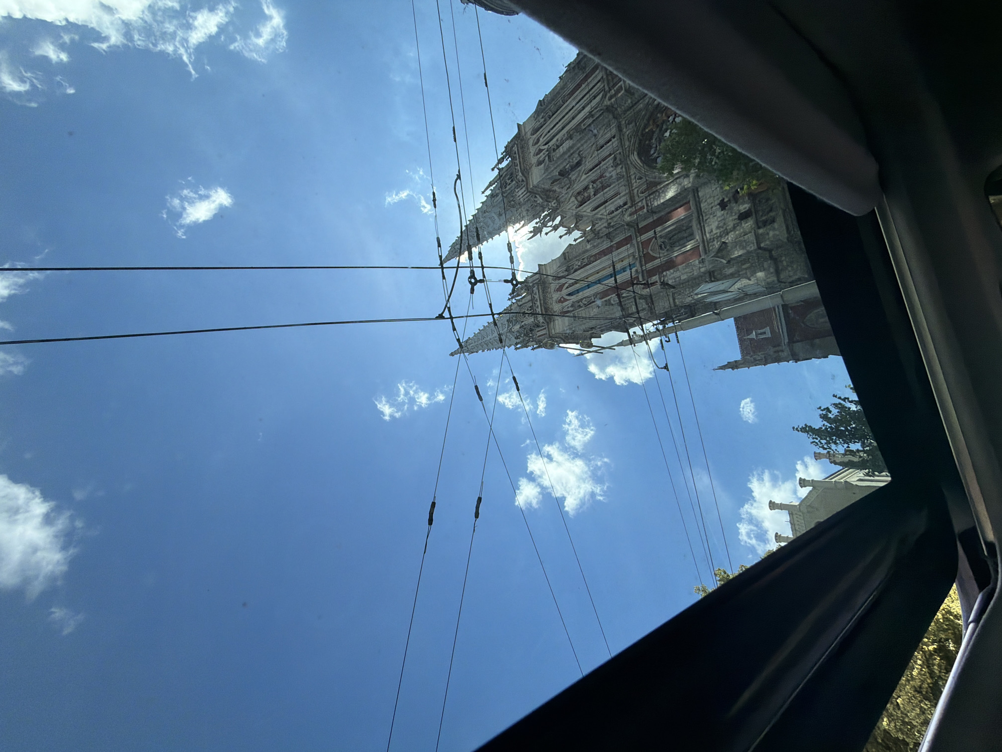 The image, taken from inside a vehicle looking up through an open sunroof, captures a low-angle view of St. Nicholas Roman Catholic Cathedral in Kyiv, Ukraine. The cathedral's neo-Gothic architecture, with its spires and intricate stonework, dominates the upper right portion of the frame. Sunlight streams from behind the cathedral, creating a bright halo effect around its towers. Overhead, a network of power lines crisscrosses the clear blue sky, which is dotted with scattered white clouds. The sunroof mechanism is visible in the foreground, framing the scene. In the lower left and right corners, parts of the vehicle's interior are visible. The surrounding environment appears to be urban, suggested by the presence of the cathedral and the overhead wires. There are no people visible in the frame.