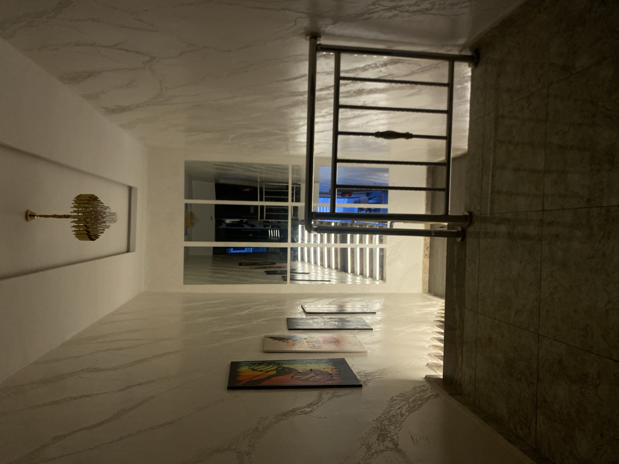 The image depicts an interior staircase with a modern aesthetic. The walls are adorned with a marble-like finish, and a rectangular ceiling fixture with a chandelier is visible overhead. Several framed artworks hang on the left wall, adding a touch of color to the neutral tones of the space. A metal railing with vertical bars lines the right side of the stairs, which appear to be made of tiled material. Through a large window at the end of the hallway, a view of another set of stairs and a dimly lit interior space is visible. The lighting in the scene is soft, with subtle illumination highlighting the textured walls and artwork. The overall impression is one of a well-lit, contemporary residential or commercial building, possibly a hotel or apartment complex. The city of Barden Yana, Nigeria is not depicted in this image.