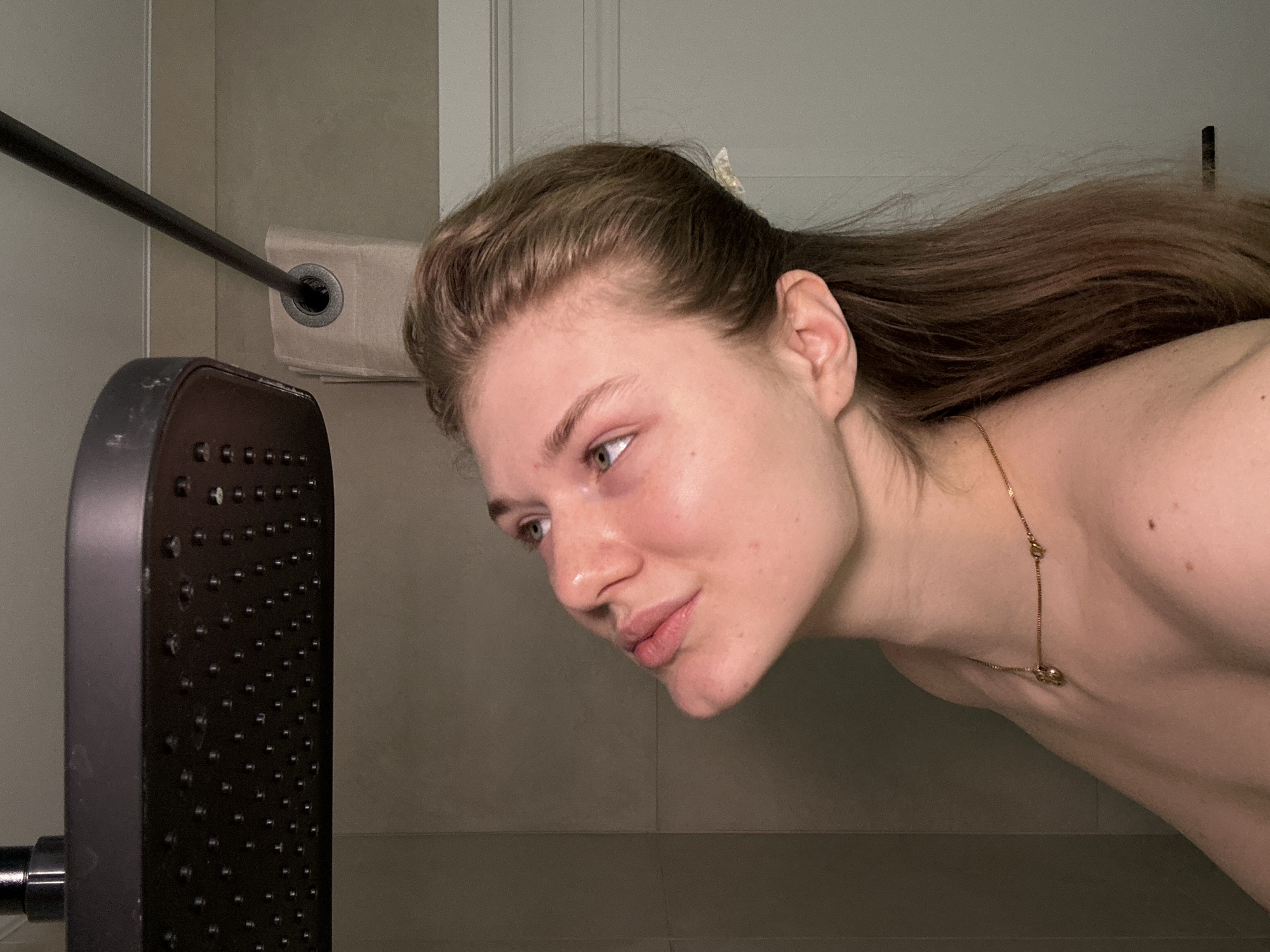 A woman is shown in a close-up shot in what appears to be a bathroom. The woman has light skin, brown hair partially pulled back with a clip, and is wearing a delicate gold necklace. She is looking intently at a shower head located to her left. The background includes a shower curtain rod, part of a beige-colored shower curtain, and neutral-toned walls. The location within the city of Kazan, Russia, cannot be determined from the image.