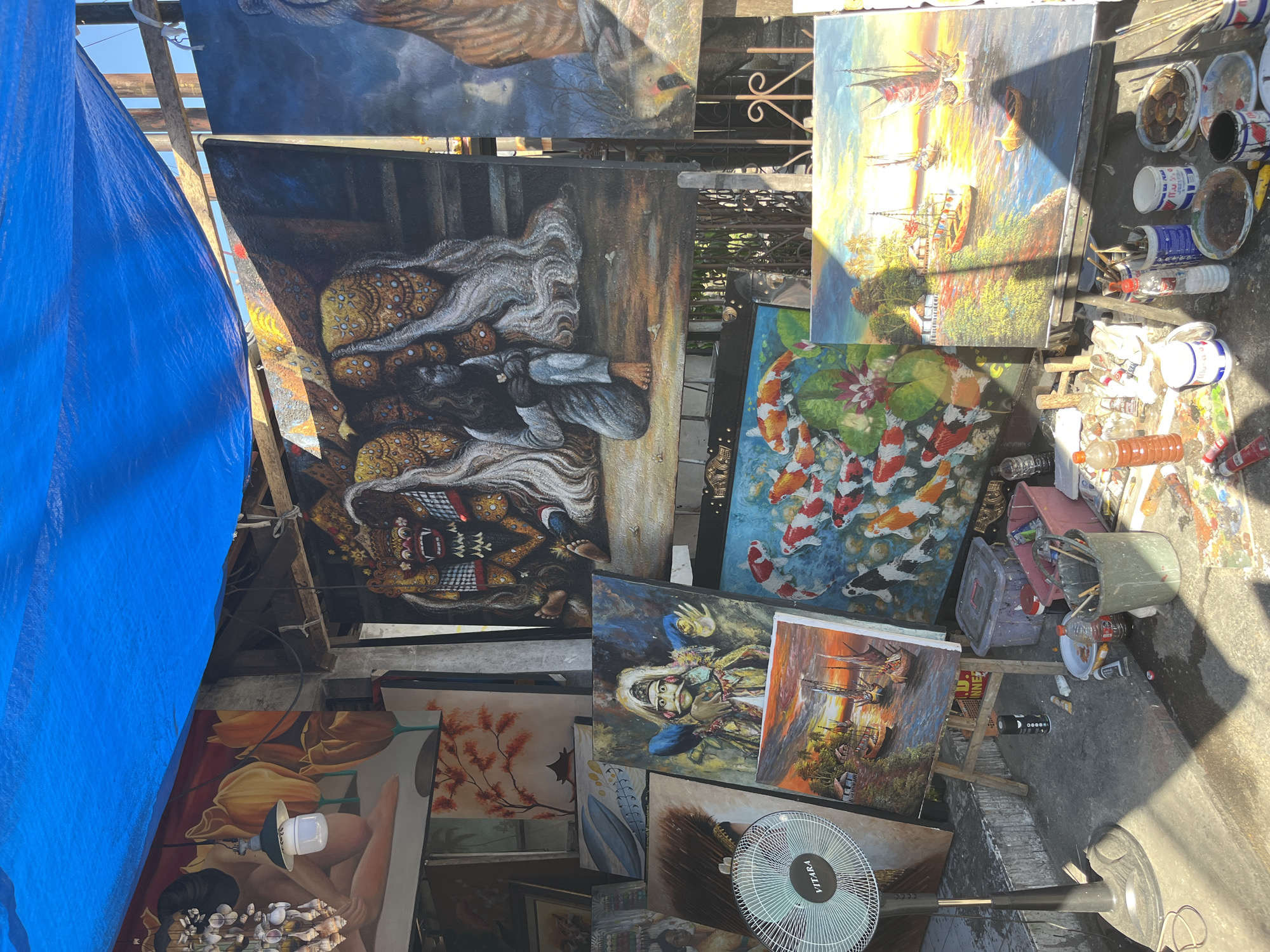 This is a medium-shot, eye-level, outdoor photograph of an art stall in Sukawati, Indonesia. A blue tarp is stretched overhead, providing shade. Numerous paintings are displayed, leaning against each other and propped on easels. The paintings feature a variety of subjects including koi fish, traditional Balinese dancers, boats sailing at sunset, and what appears to be a Balinese demon. A fan is positioned on the left side of the frame. Various art supplies, such as paint tubes, brushes, and containers, are scattered around the base of the easels and on the ground, suggesting an active workspace. The overall impression is that of a vibrant, informal art market.