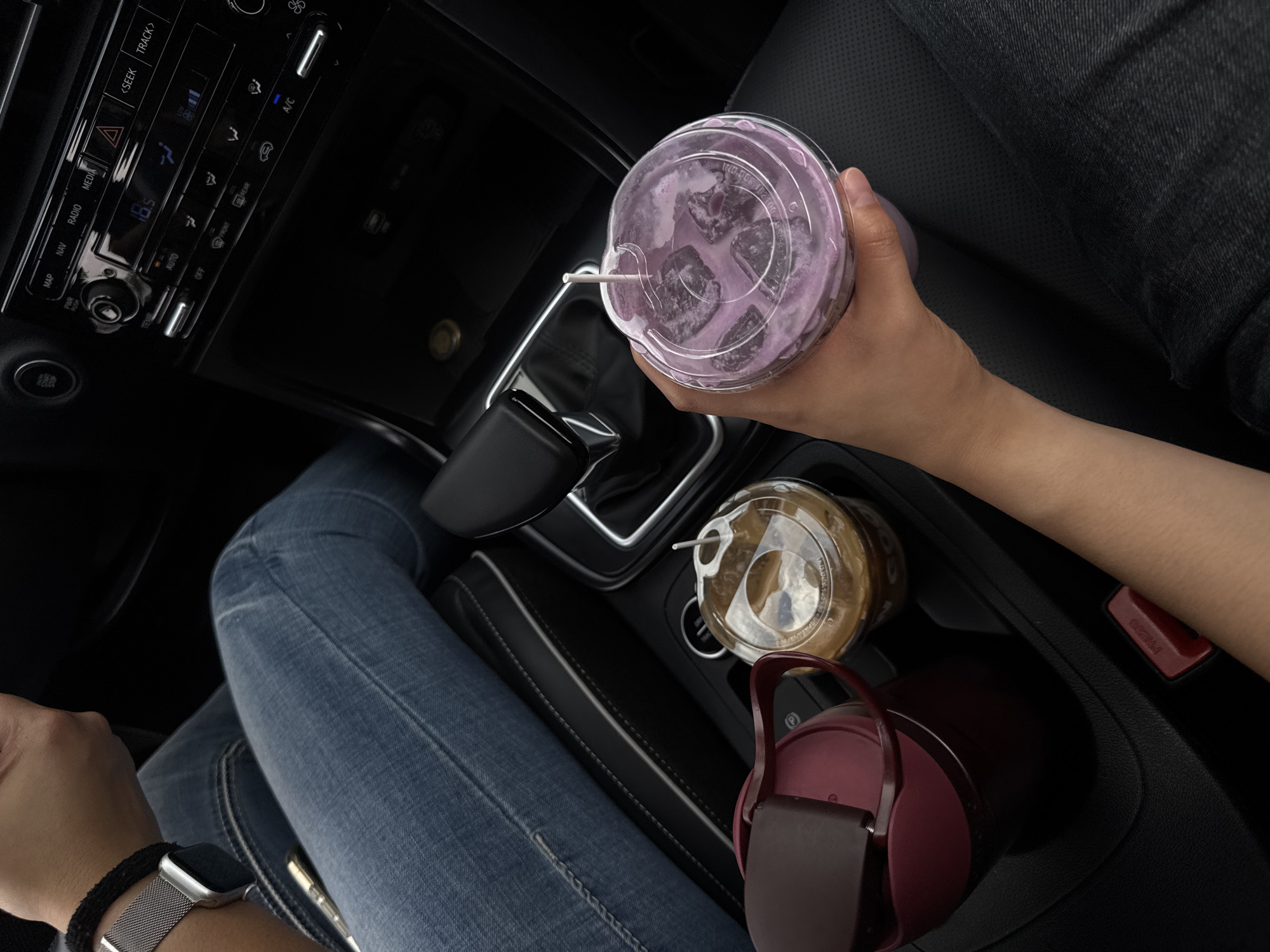 A high-angle, top-down view captures the interior of a car, focusing on a person's body parts and multiple beverages. A person, dressed in blue jeans, is seated, with their left arm visible. A silver smartwatch with a mesh band and a dark wristband adorns their wrist. The car's interior is dark, featuring a center console with a gear shifter and cup holders.
In the cup holders, a clear plastic cup containing a light brown iced drink is visible, alongside a dark red water bottle. Another arm, entering the frame from the right, holds a clear plastic cup filled with a purple iced beverage, complete with a straw and visible ice cubes. The drink has a creamy, swirled appearance.
The car's dashboard is partially visible on the left, displaying various buttons and a digital screen showing "18.5," likely a temperature reading. Visible text on the dashboard includes "MAP", "NAV", "RADIO", "MEDIA", "<SEEK", "TRACK>", "PWR PUSH", "AUTO", "OFF", "FRONT", "REAR", and "A/C". On the lid of the purple drink cup, part of the text "IND.DES 102100" is discernible. A red latch near the cup holders features the word "PRESS". The scene depicts a casual moment inside a vehicle, potentially in Puebla, Mexico.