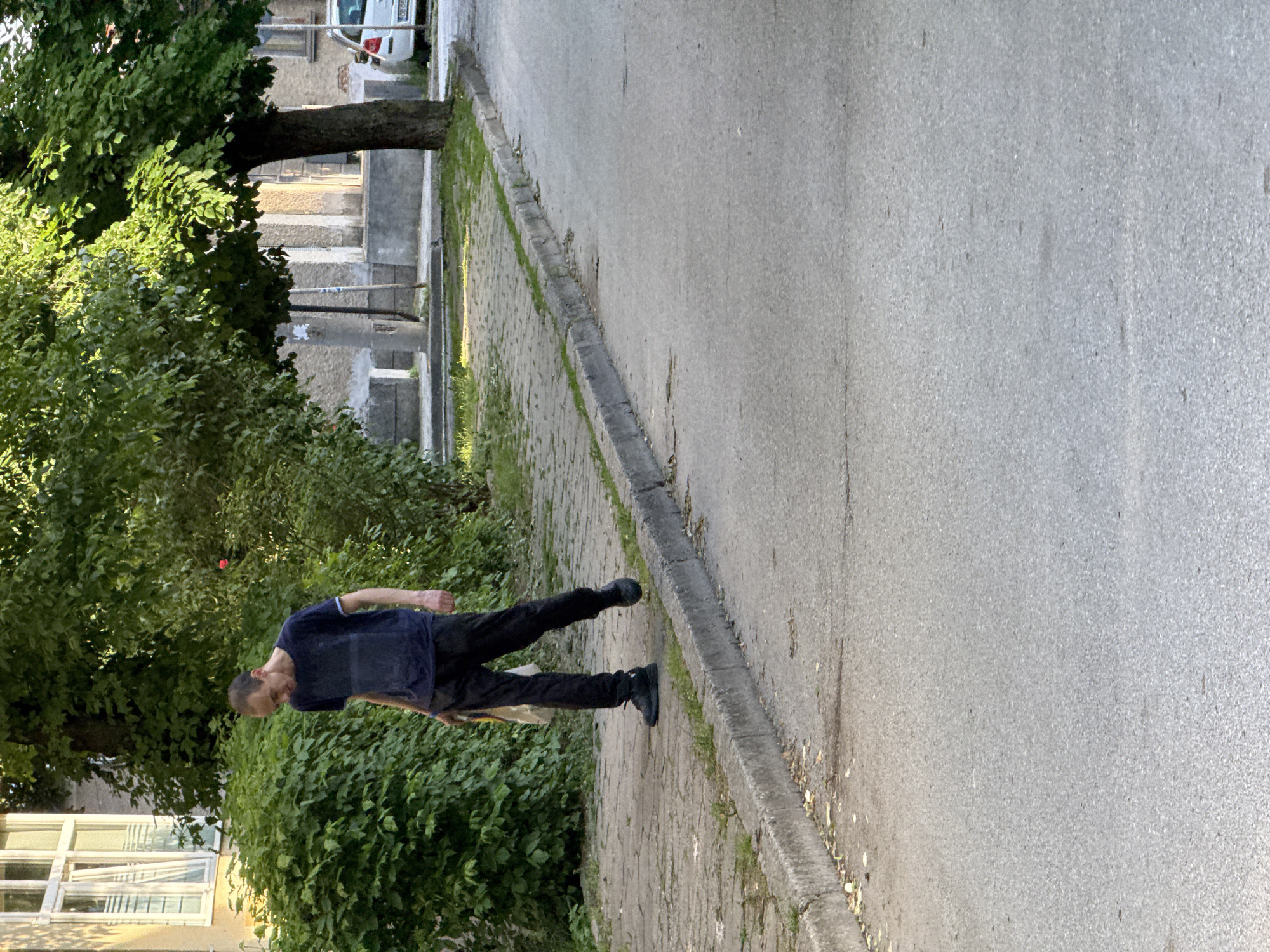 The image shows a man walking on a paved sidewalk next to a street. The man is wearing a dark blue t-shirt and dark pants, and dark shoes. To the left of the sidewalk is dense green vegetation and trees. In the background, there is a building and a white car. The location appears to be an urban area. The location is Lovech, Bulgaria.