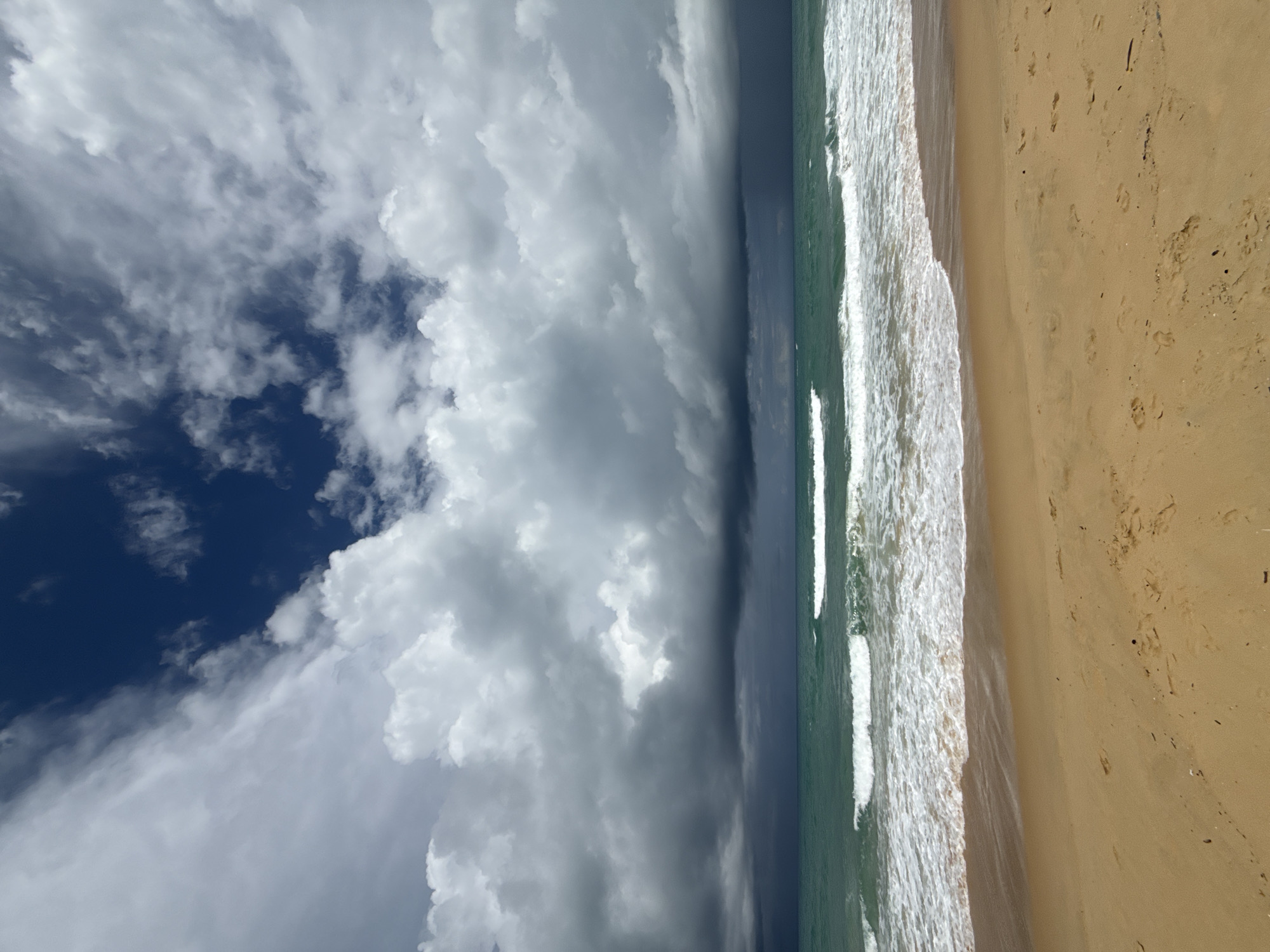 This image depicts a wide, sandy beach at Karon, Thailand, under a dramatic sky. The foreground is a tan-colored beach with a few scattered footprints. The middle ground is the ocean, with turquoise water and white-capped waves rolling towards the shore. The horizon is marked by a distinct line of dark, stormy clouds, with lighter, puffier clouds above them. The sky transitions from dark gray and blue near the horizon to a brighter blue with white clouds further up. There are no people or man-made objects visible in the scene.
