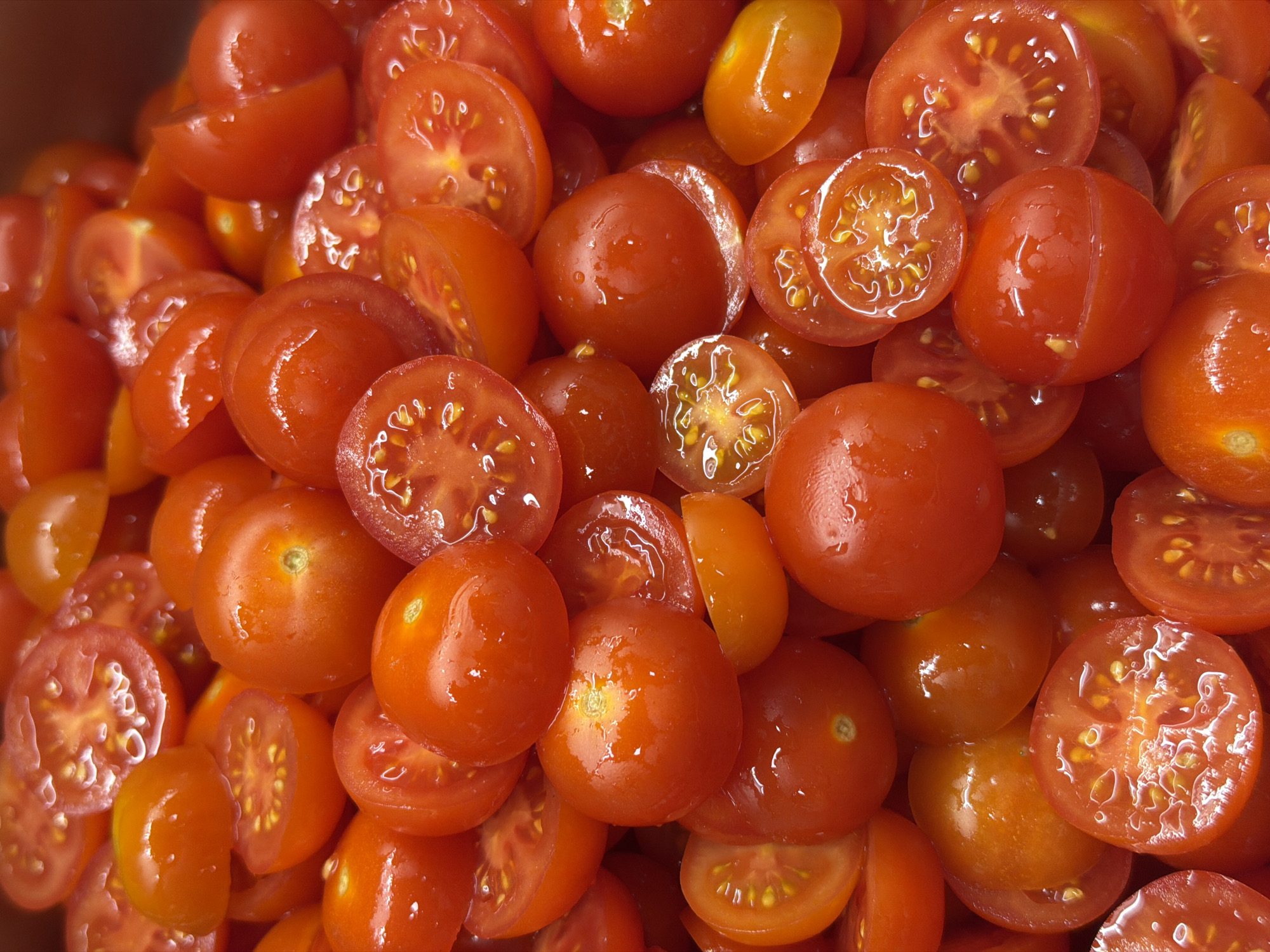 The image shows a close-up of many ripe cherry tomatoes. Some are whole, while others are sliced in half. The cut tomatoes reveal their inner structure with seeds. The tomatoes have a shiny appearance, possibly due to moisture or a coating. The scene has no indication of being in Belgrade, Serbia.