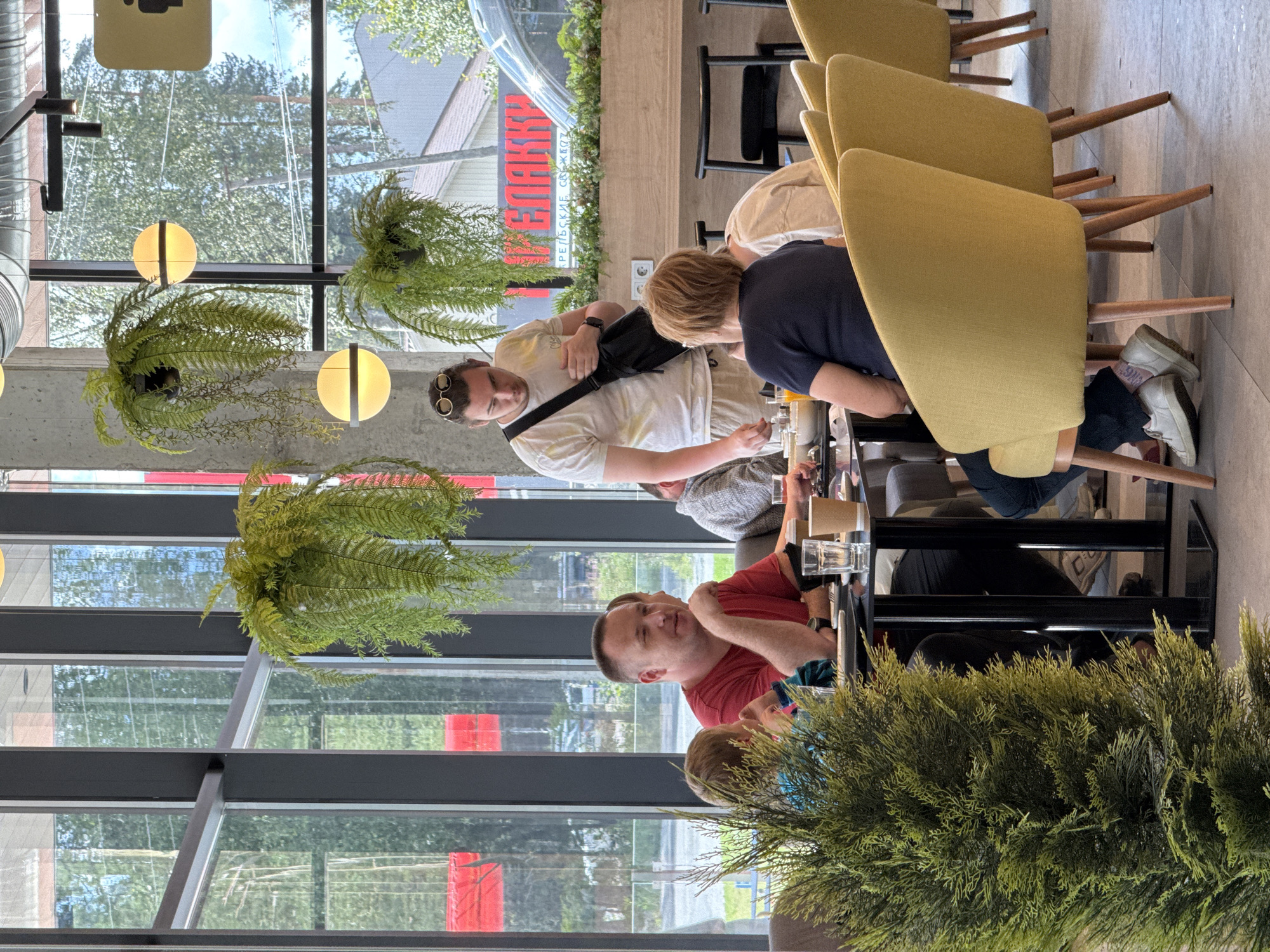 A group of people are seated at a table in a bright, modern cafe with large windows overlooking a street in Lakhdenpokhya, Russia. A man in a red shirt is talking to a child, while a young man stands behind the table with a bag slung over his shoulder. Other individuals are seated around the table, and there are plants hanging from the ceiling and a potted tree in the foreground. The cafe appears to be in a commercial area, with buildings visible through the windows.