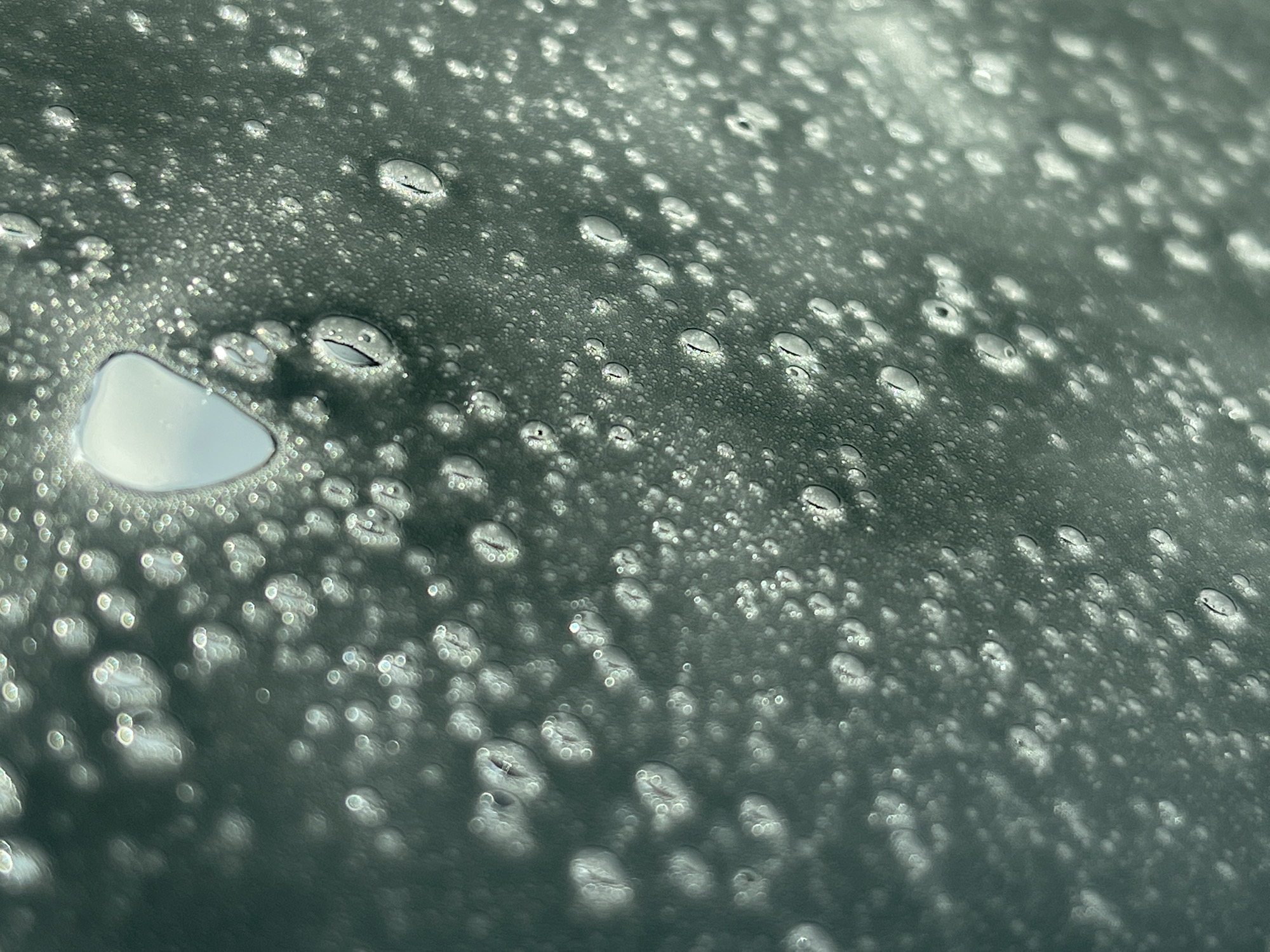 The image shows a close-up view of a surface covered in water droplets. The surface appears to be a light green or grey color. The droplets vary in size, with some being quite large and others very small. The large droplet at the left side of the image is translucent and reflects the environment with higher definition. The environment where the image was taken is not visible. There is no information about the location being the city of Chaikovsky, Russia, or about people, actions, interactions, or specific objects in the image.