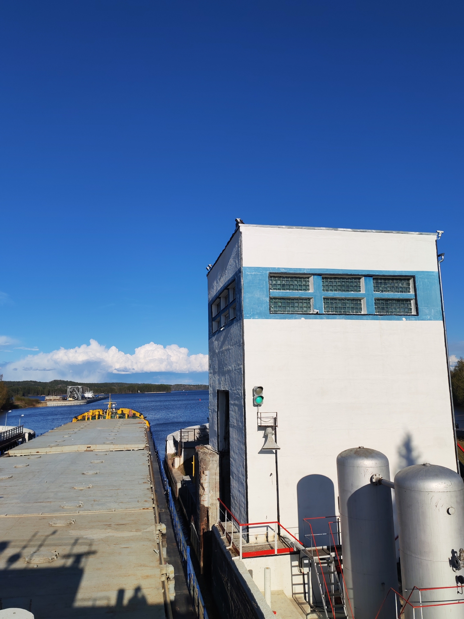 A medium-angle, outdoor shot captures a barge docked beside a white building with a blue stripe near Белоусово, Russia. The barge, a gray, flat-topped vessel, sits between the building and a dock or riverbank, partially obscuring the view of the water. A structure is visible in the distance on a body of water, appearing under a bright blue sky with scattered clouds. The building adjacent to the barge features a row of small, glass-block windows within the blue stripe. Below the stripe, the building is primarily white, with various industrial fixtures, including cylindrical tanks, a red-and-white staircase, and a traffic signal showing a green light. The lighting in the shot suggests a clear day, with shadows cast by the building and tanks.