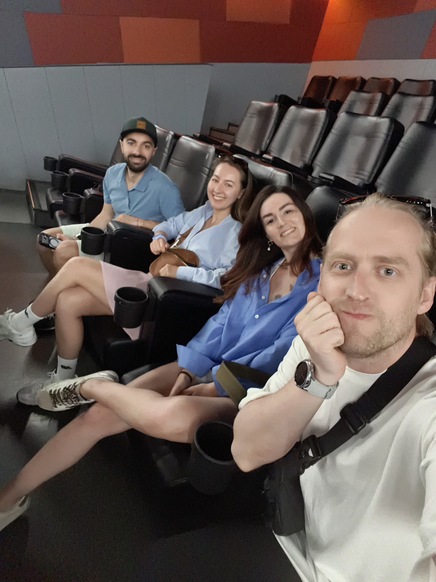 Four individuals are seated in a row of cinema chairs. The person in the foreground, on the right, is a white male with blond hair, wearing a white long-sleeved shirt and a watch on his left wrist. He is looking at the camera and resting his chin on his hand. To his left, a white female with long brown hair is seated, wearing a blue collared shirt. Further to the left, another white female with long brown hair, wearing a light blue collared shirt and a pink skirt, is visible. She is holding a brown bag. The leftmost individual is a white male with dark hair and a beard, wearing a blue collared shirt and a baseball cap. He is holding a black object in his right hand. The chairs are black, and cup holders are visible between them. The background appears to be a wall with gray and orange geometric shapes. The setting is consistent with a cinema or screening room. This scene is not located in Tyumen, Russia.