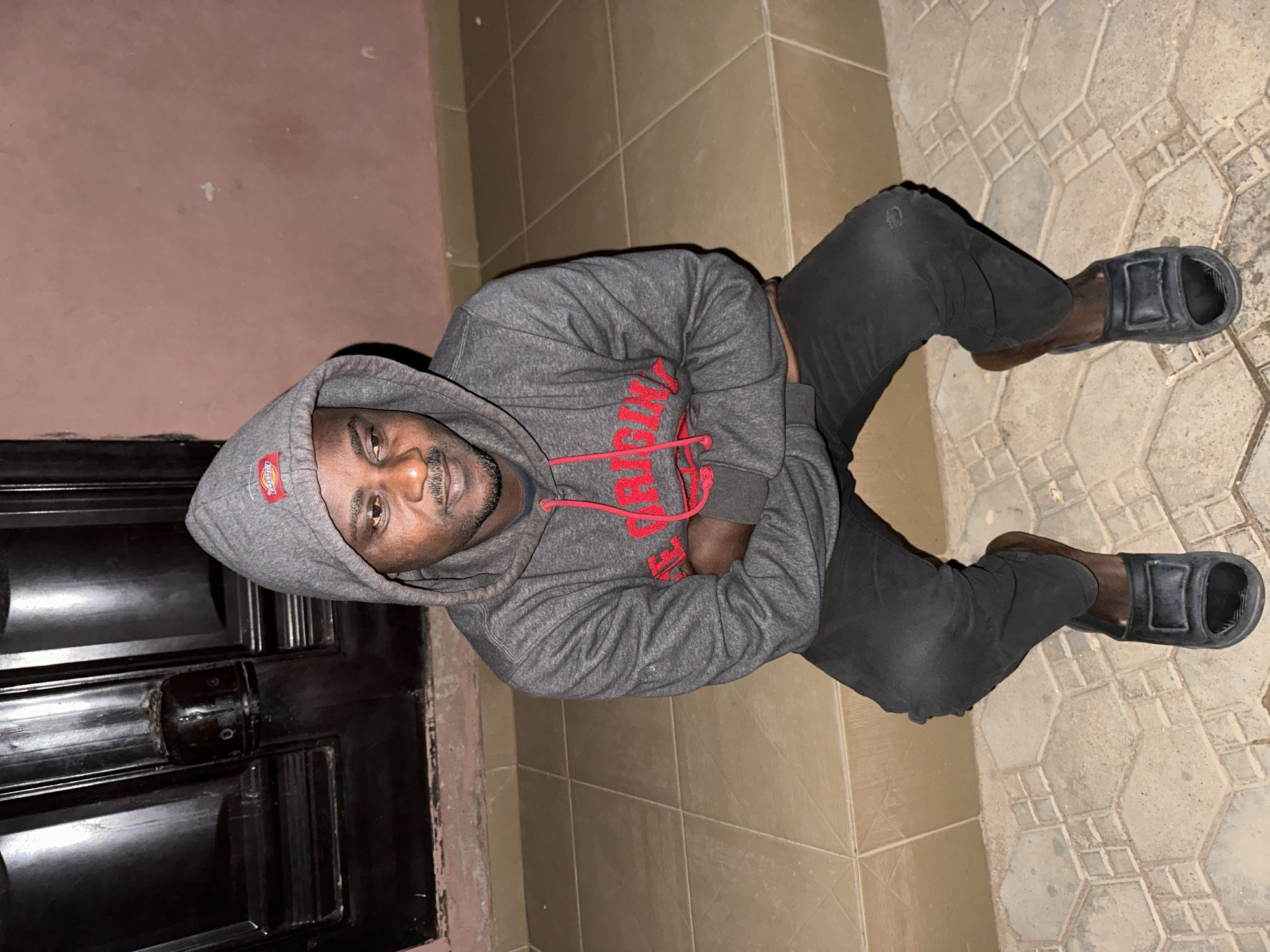 In Narayi, Nigeria, a Black man wearing a grey hooded sweatshirt with red drawstrings and the words "ORIGINAL" in red lettering, and black pants, sits with his arms crossed. He is wearing black open-toed sandals with brown socks. He is positioned on a light brown tiled step with a darker tiled walkway in front of him. Behind him is a dark wooden door with a textured surface and a dark pink wall. The lighting suggests it is nighttime.