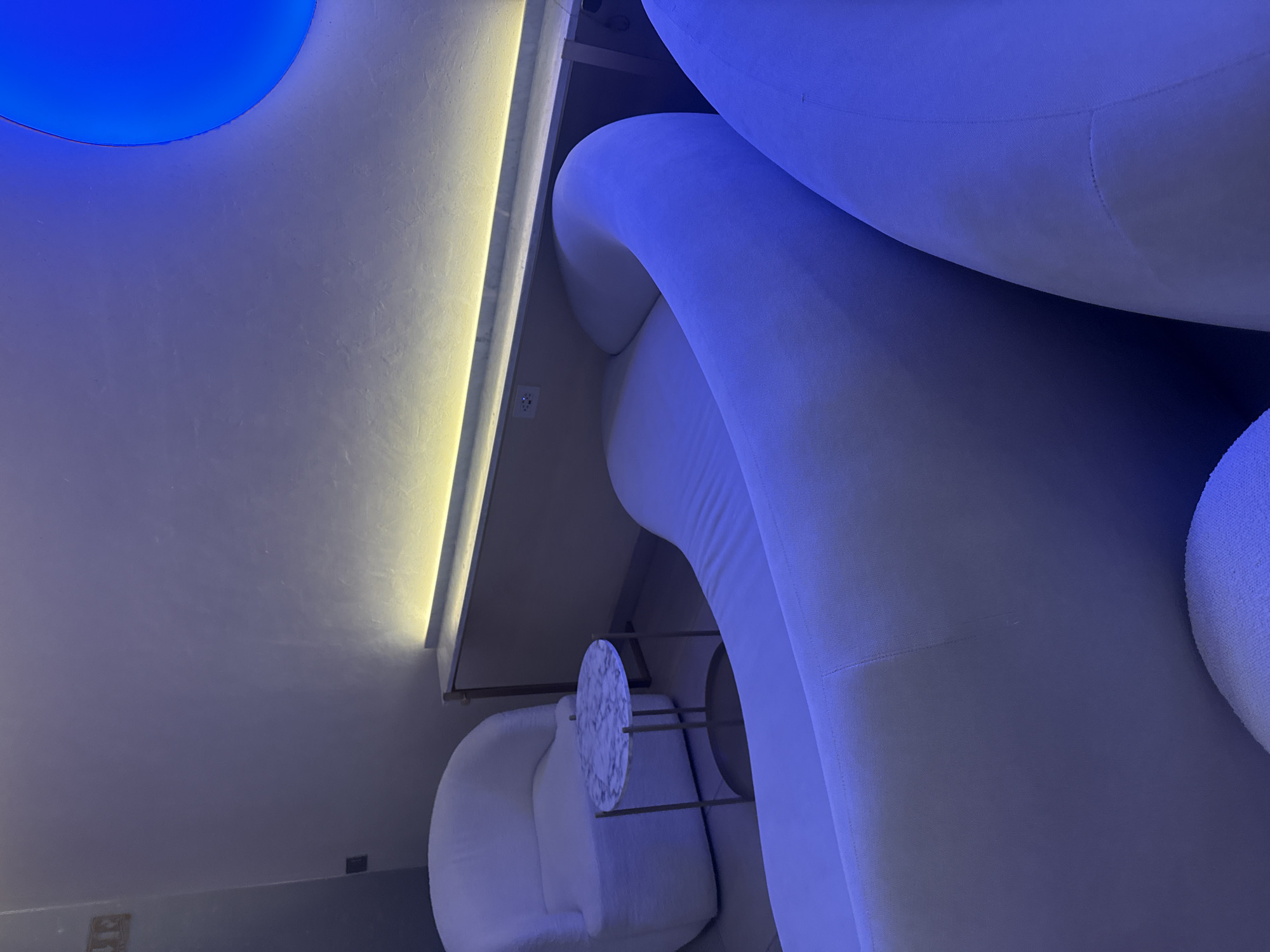 The image captures an interior space with contemporary design elements, likely located in San Bernardino Tlaxcalancingo, Mexico. The dominant features are plush, rounded white sofas, partially illuminated with a blue hue. A small, round marble-topped table with a metal frame sits near a white armchair. The ceiling has a recessed lighting design, featuring both a blue circular light and a linear yellow light. The walls are a neutral color. The overall impression is that of a modern, stylized lounge or waiting area.