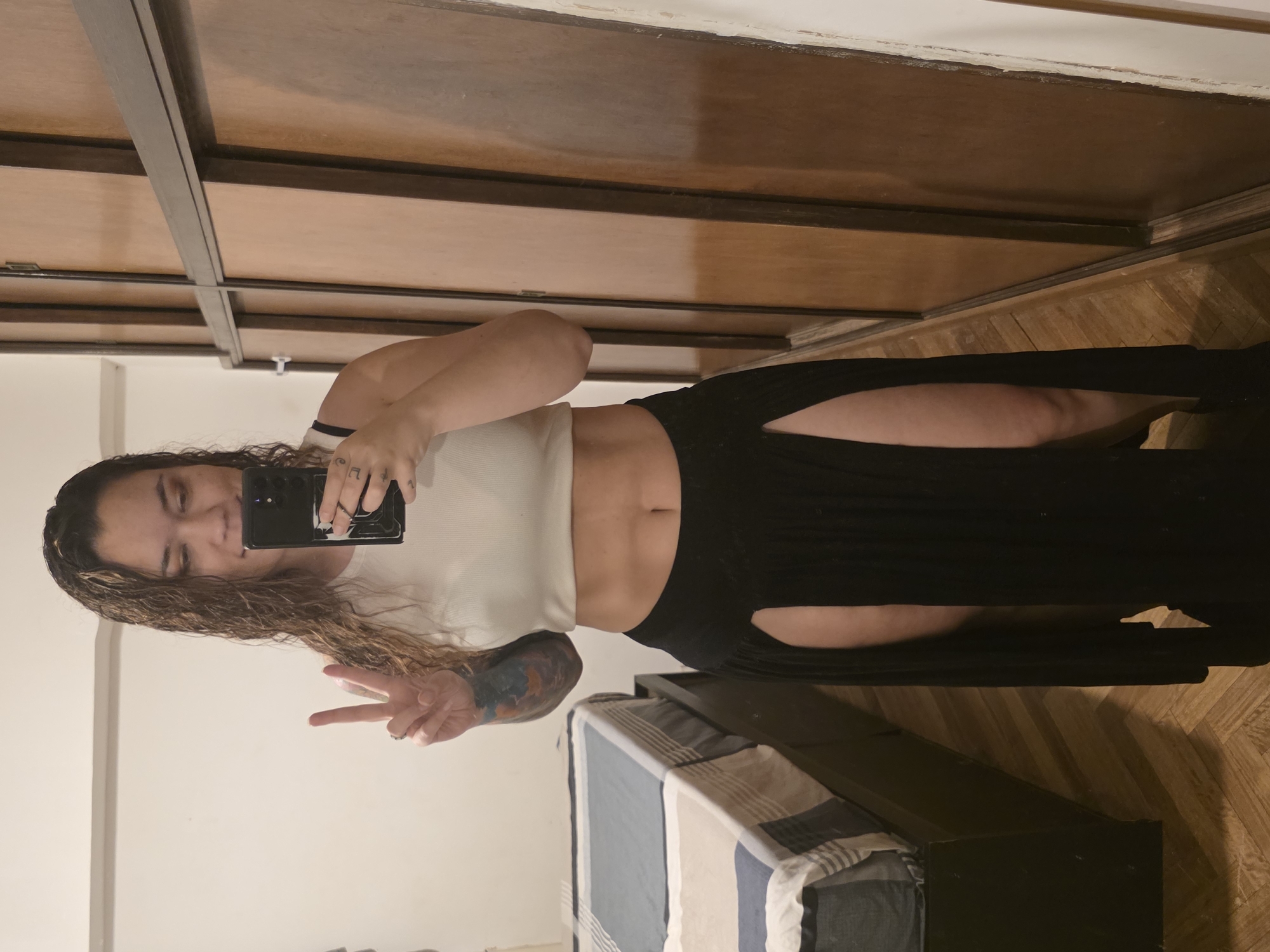 A female individual is depicted in a full-length mirror selfie within an indoor domestic setting, consistent with residential architecture found in Buenos Aires, Argentina. The subject, with long, wavy, damp-appearing brown hair, is wearing a white ribbed crop top exposing the midriff, and a black maxi skirt with high side slits. A colorful full sleeve tattoo is visible on her left arm, and a small dark tattoo is present on a finger of her right hand. She holds a black smartphone in her right hand to capture the image, while her left hand is raised, forming a 'peace' sign. Her facial expression shows a slight smile. The background on the left features a bed with a dark frame and a striped blue, white, and beige comforter. On the right, a large wooden wardrobe with multiple panels and dark framing is visible. The floor is covered with light-colored herringbone-patterned wood, and the walls are white.